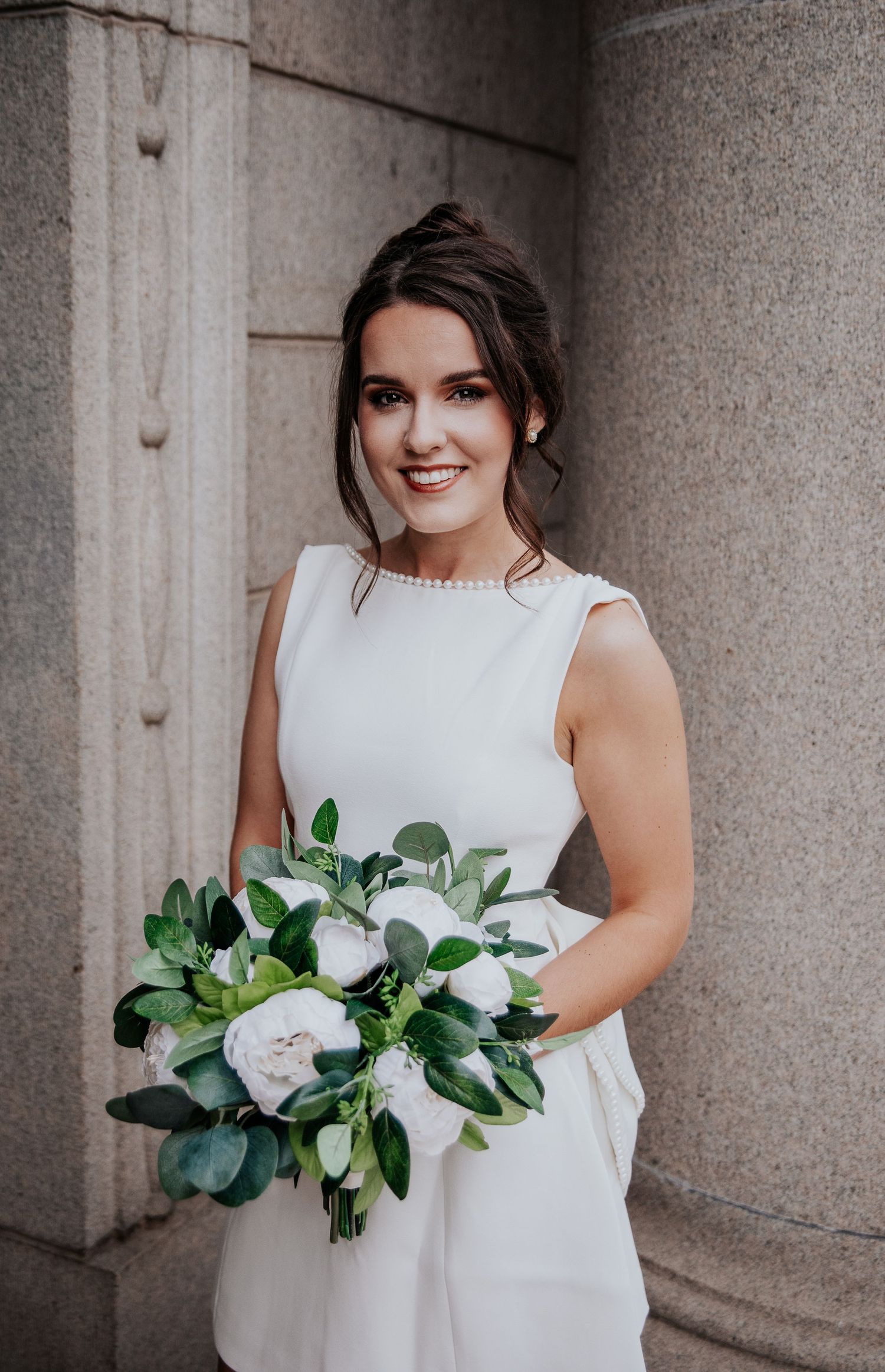 A bride in a white sleeveless dress holds an elegant bouquet of white flowers and greenery against a stone column backdrop .