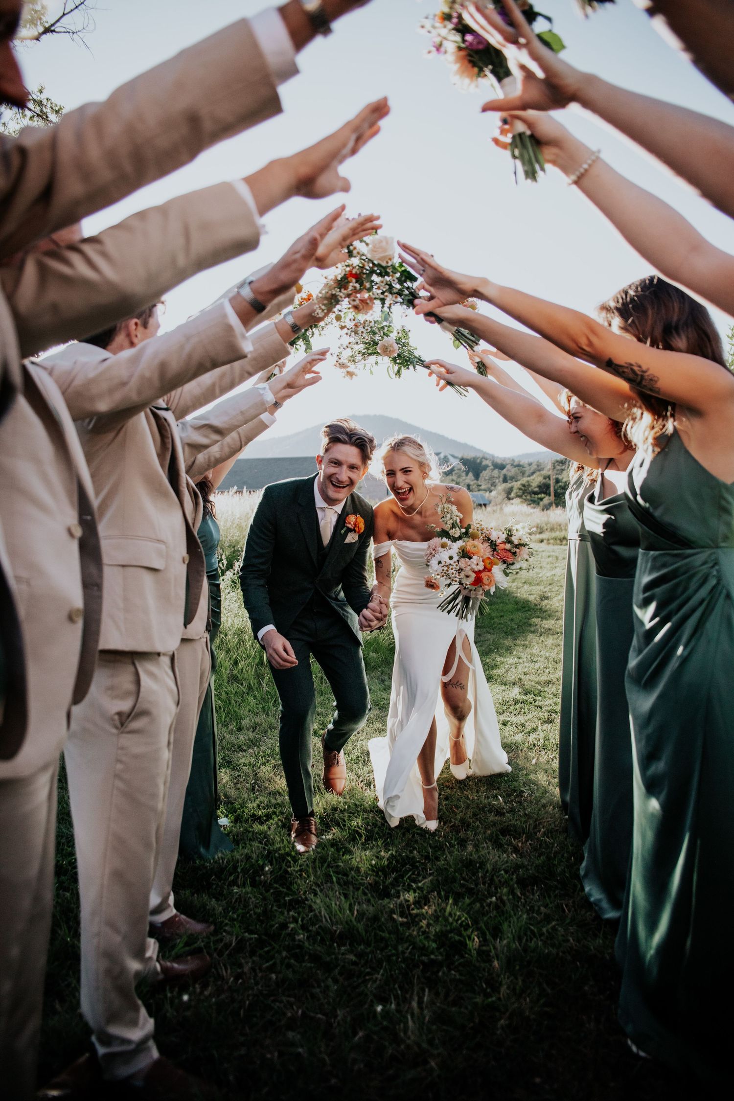 Wedding party celebrates with raised hands and flower petals on a scenic hillside during outdoor ceremony.