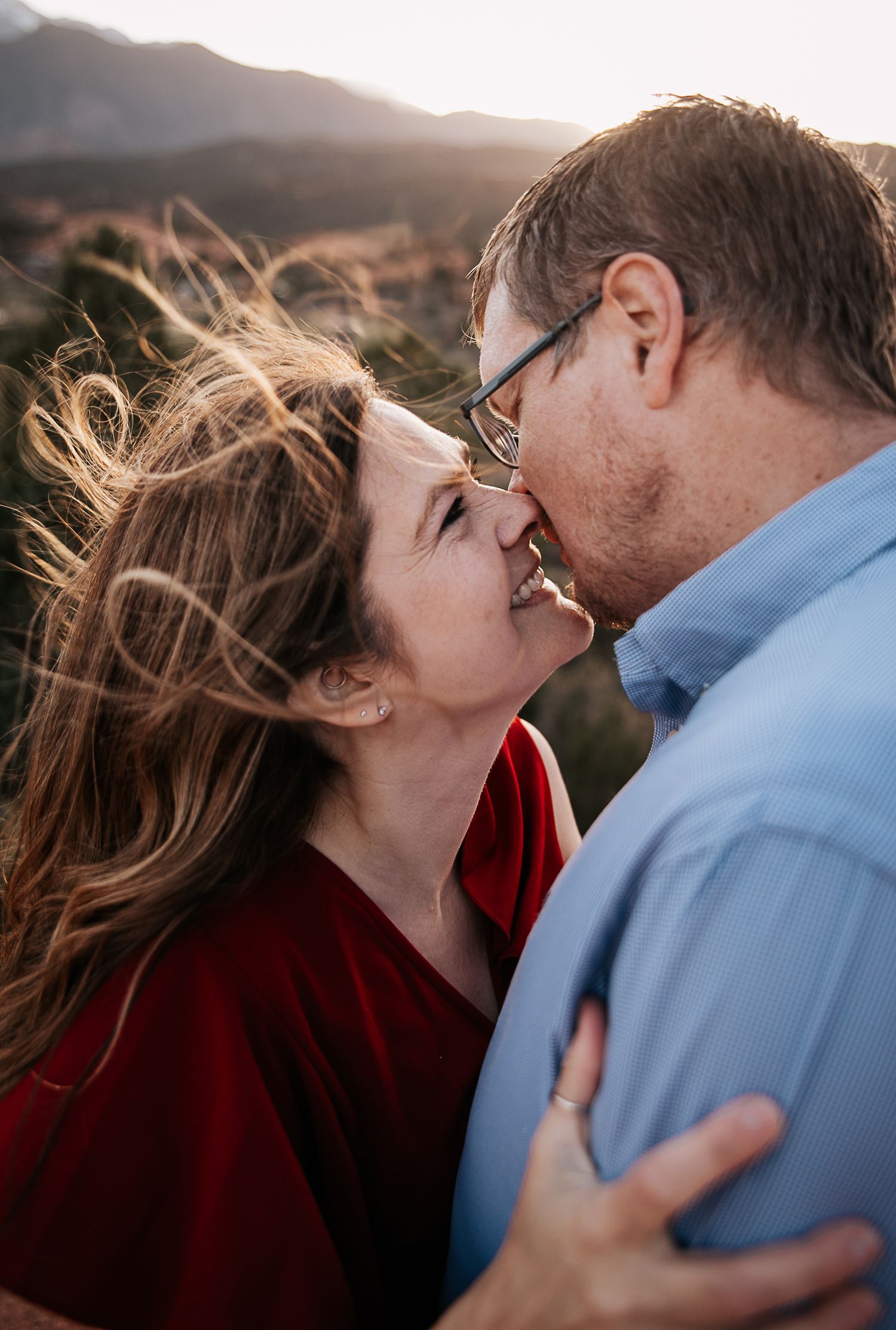 Wind-swept couple shares an intimate moment against mountain backdrop during golden hour.