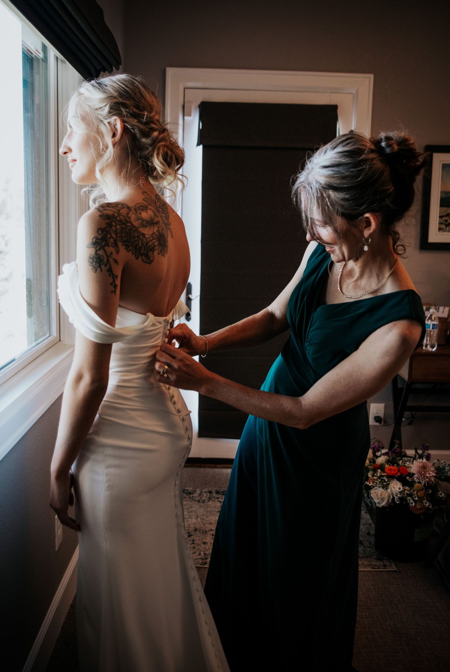 Helper in teal dress assisting with a fitted white wedding gown near a window.