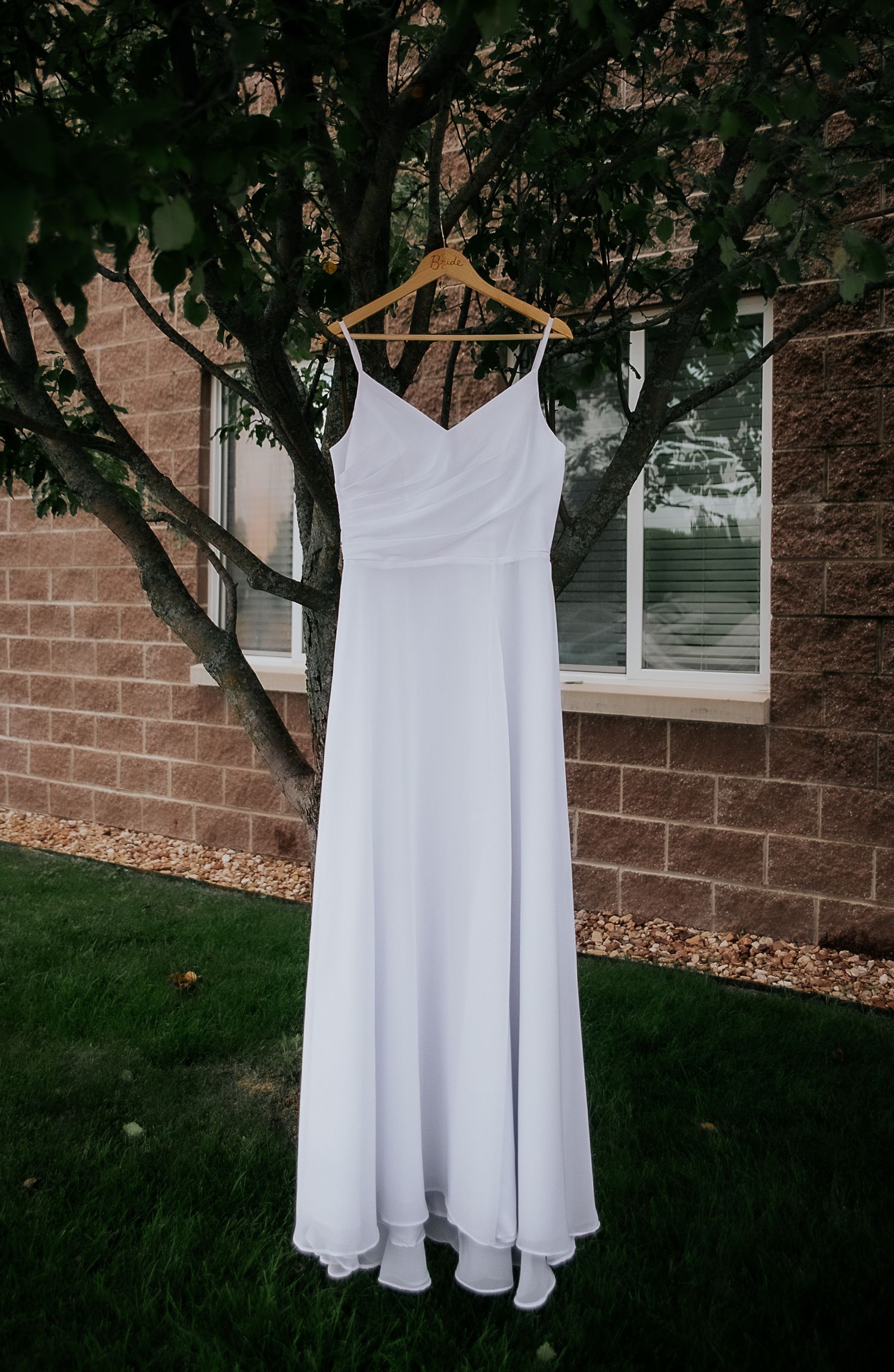 Elegant white wedding dress hanging on wooden hanger against brick wall with green foliage.