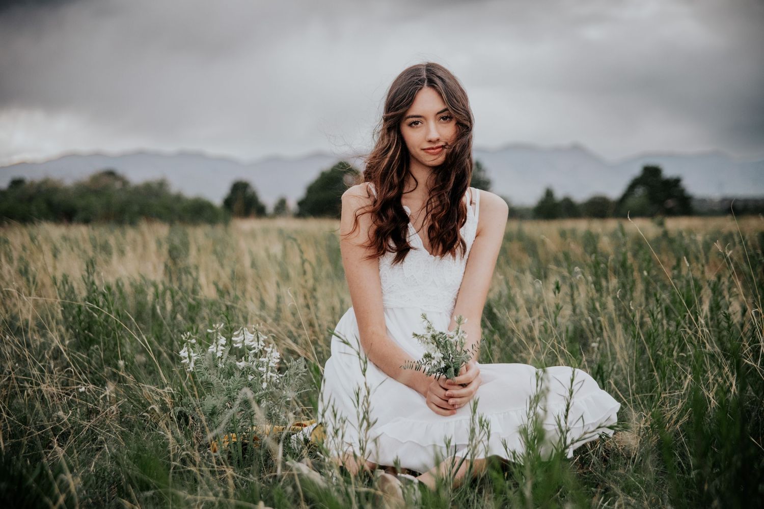 A person in a white dress sitting gracefully in a grassy meadow with mountains in the background.