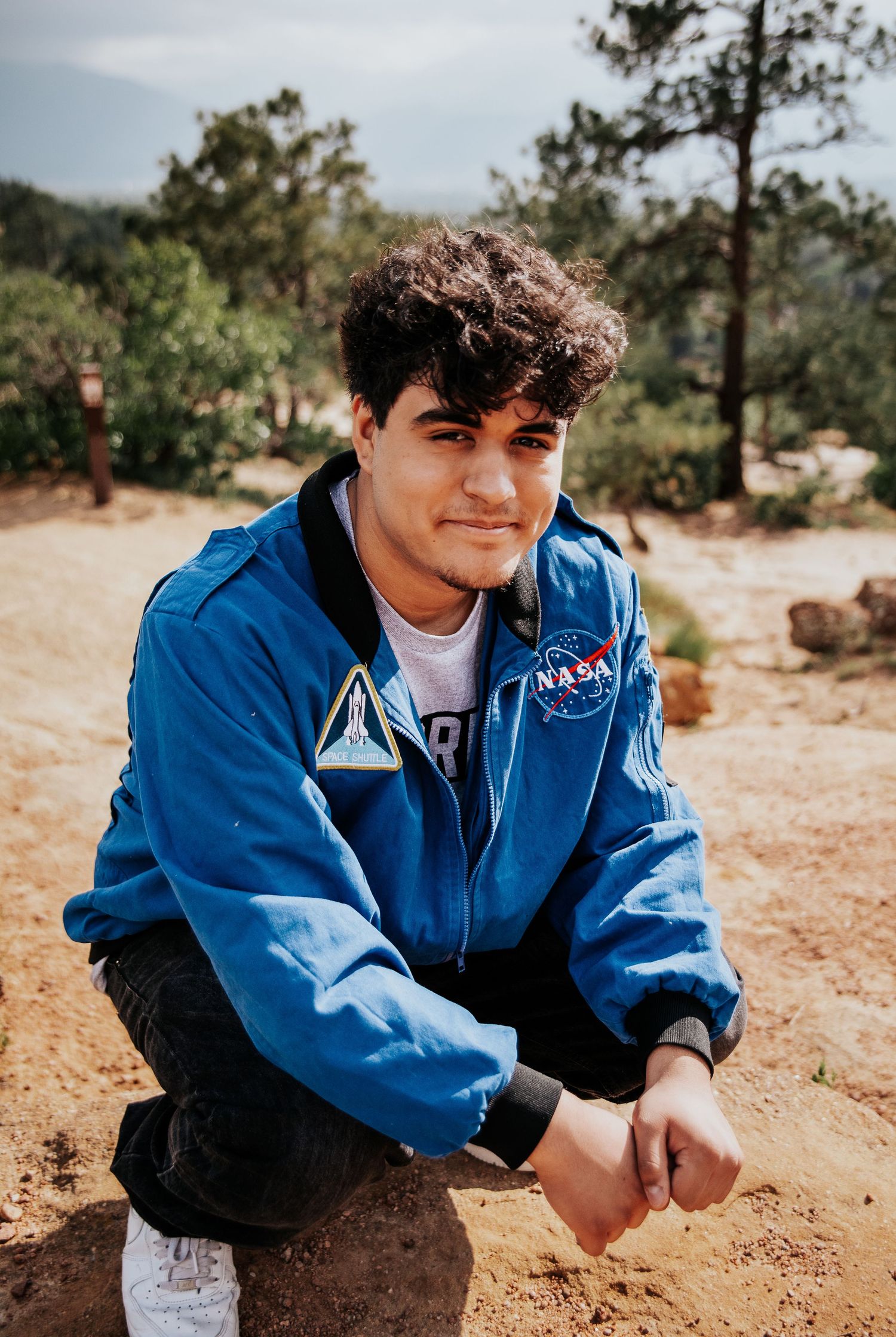 High school senior in blue NASA jacket sits on dirt ground at Palmer Park in Colorado Springs