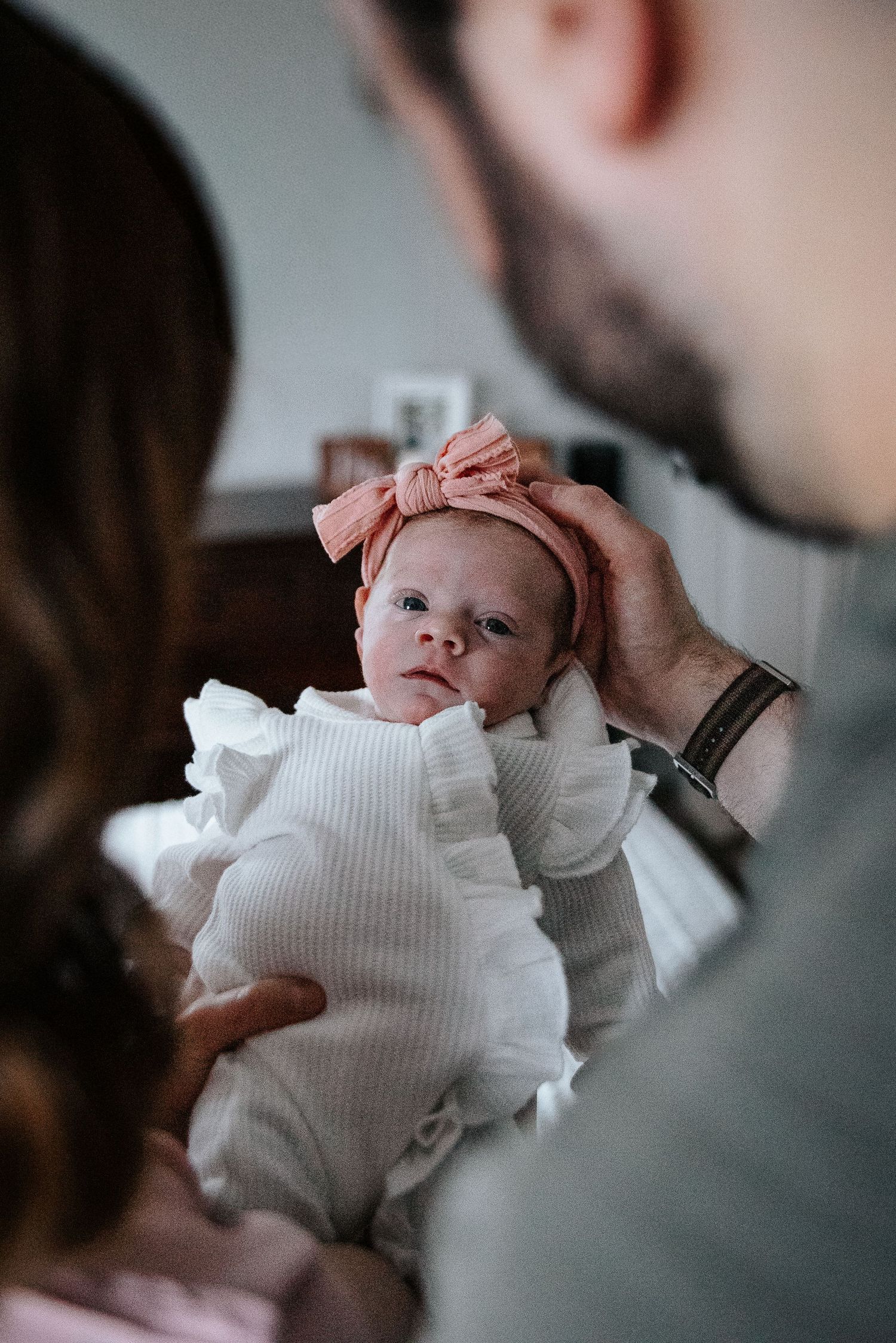 A newborn baby wearing a white ribbed outfit and pink bow is held tenderly during an intimate family photo session.