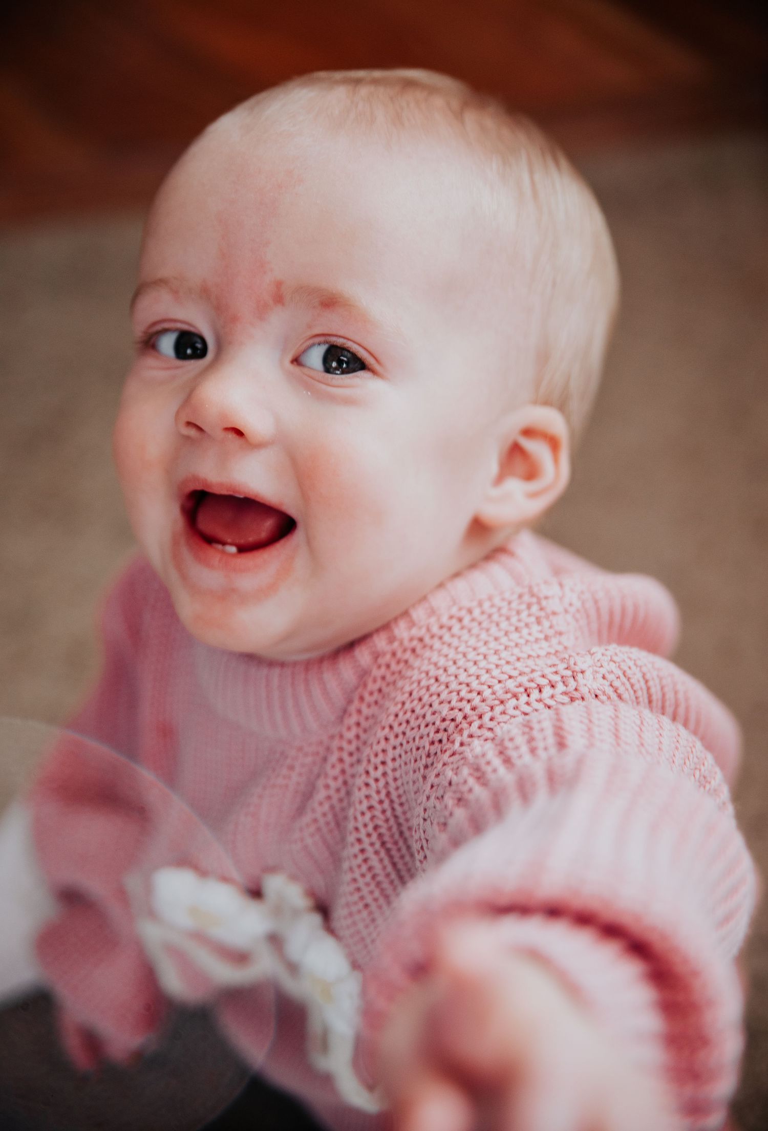 Baby wearing pink knit sweater smiles while laying on blanket.