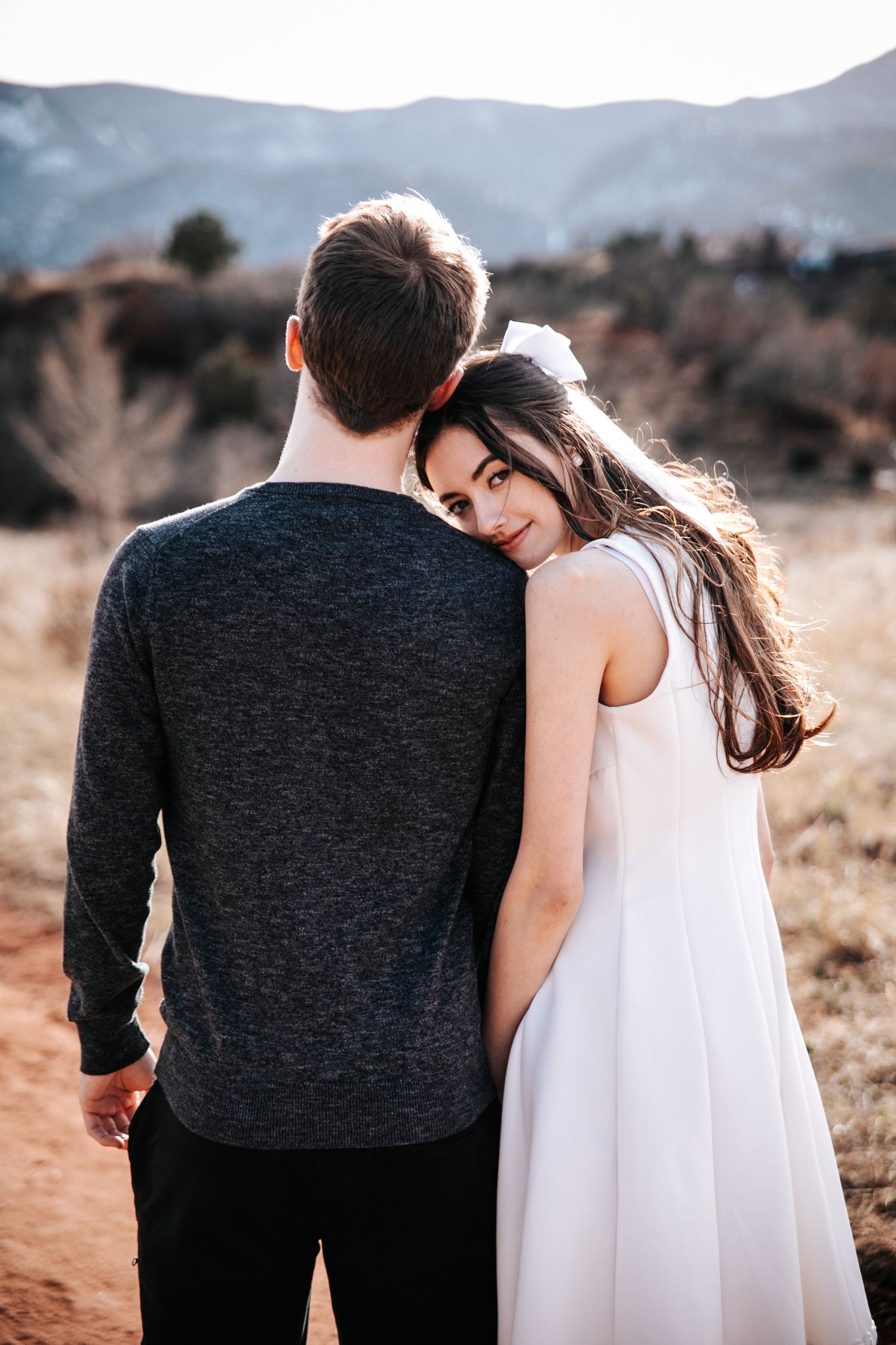 Back view of couple standing together against mountain landscape with woman in white dress.