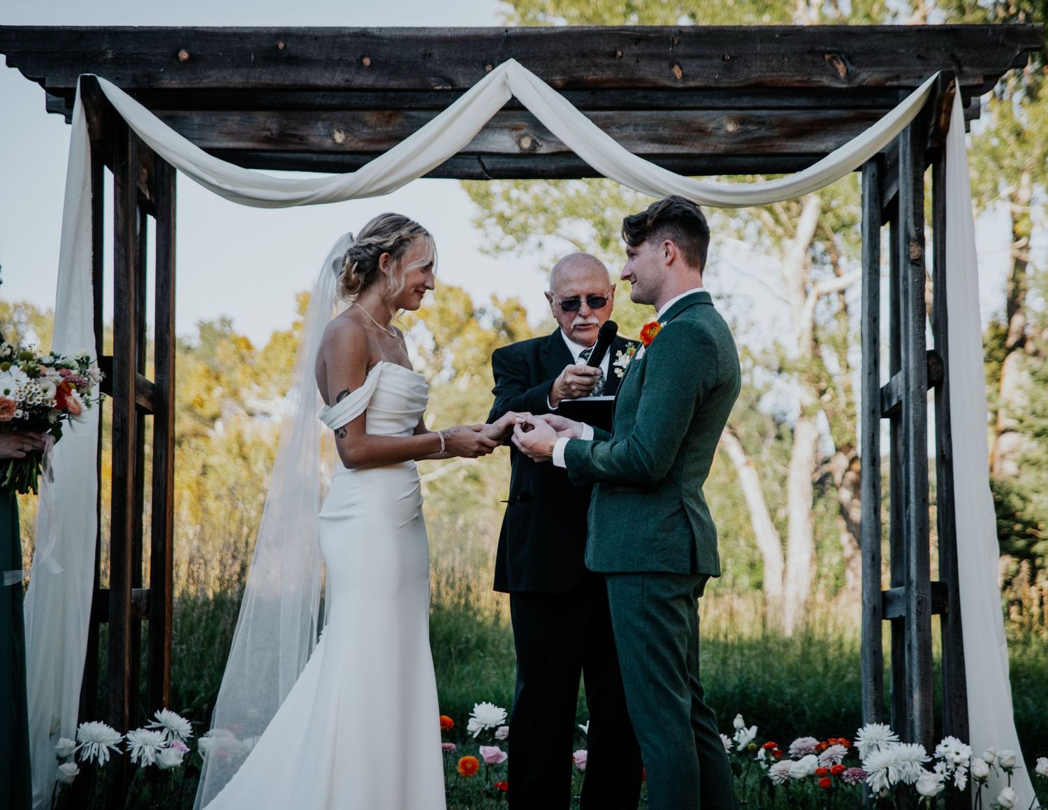 A couple exchanges rings during their wedding ceremony under a decorated wooden arch at an outdoor venue.
