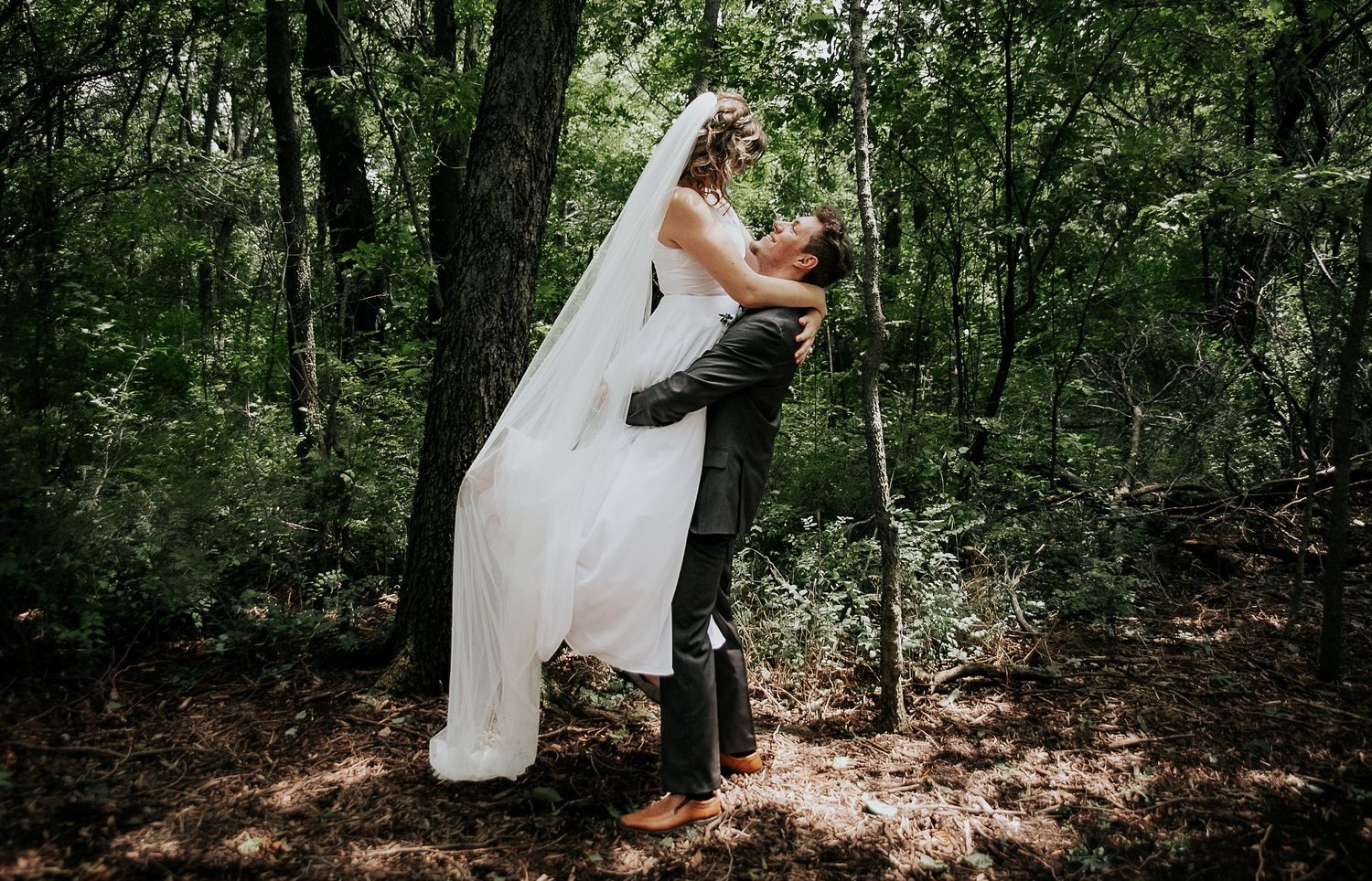 Couple embraces in romantic pose with flowing veil in wooded setting.