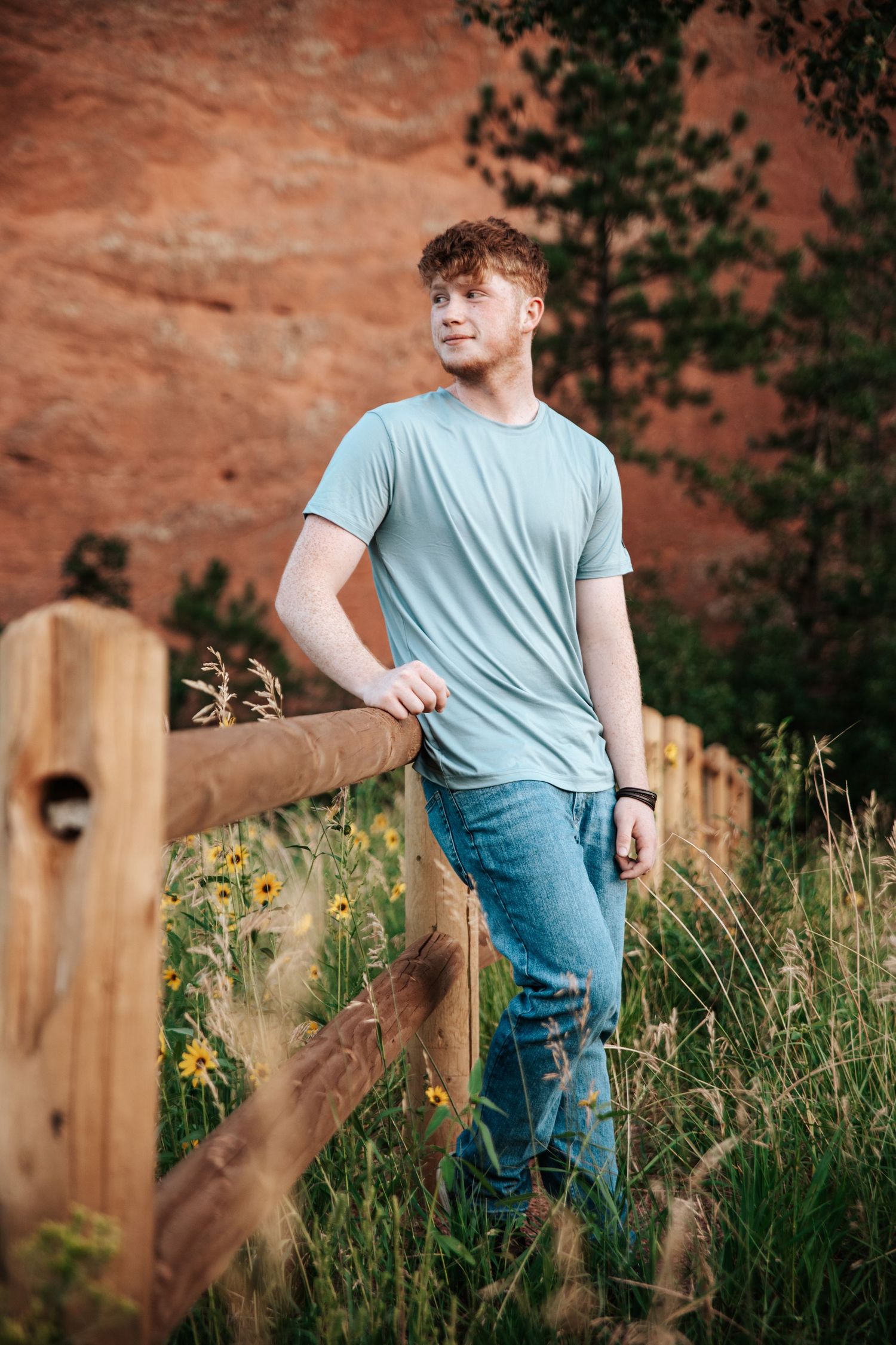 Person in light blue shirt leans on wooden fence with red rocks and yellow wildflowers in background.