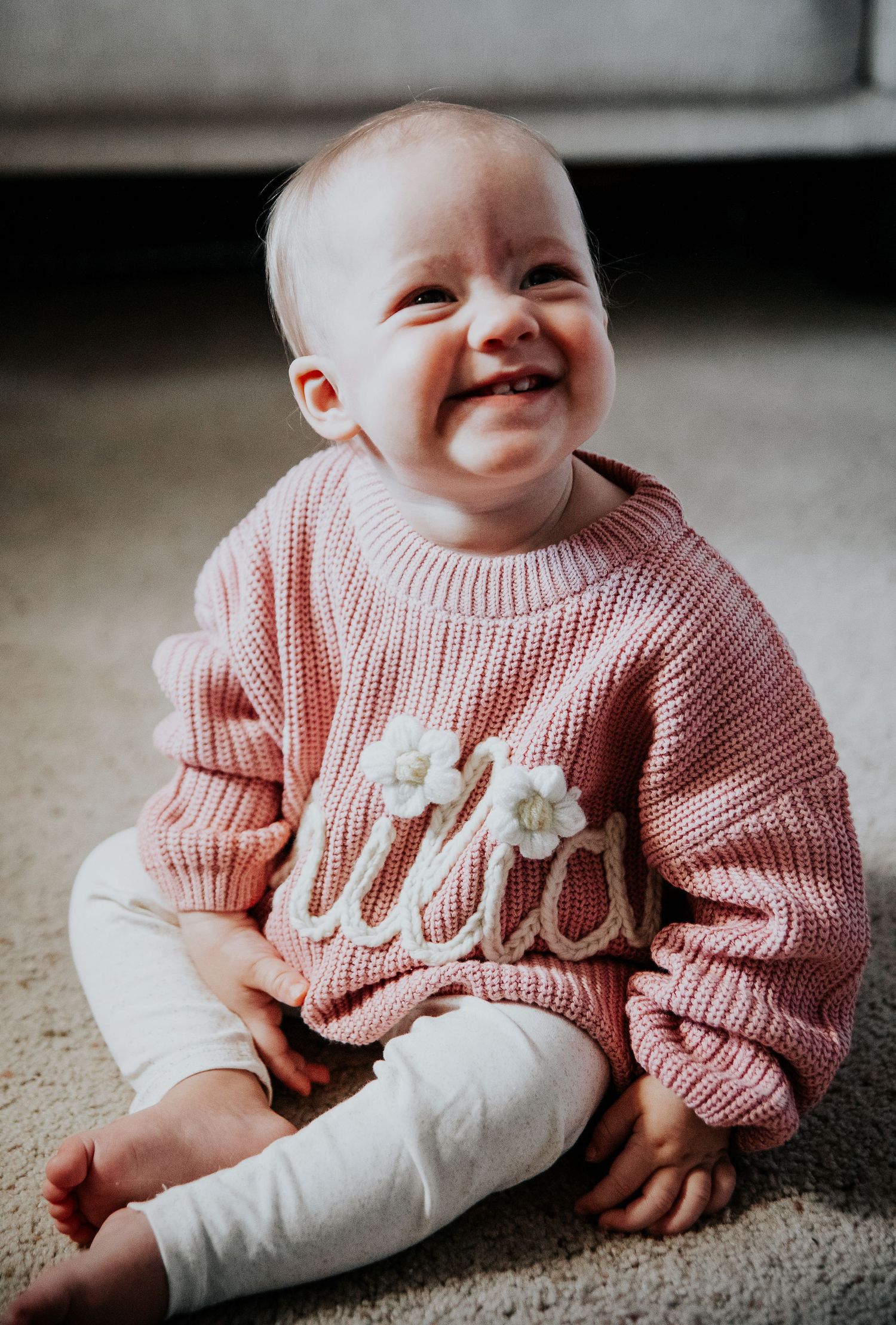 Baby in pink sweater sits on floor showing joyful expressions.