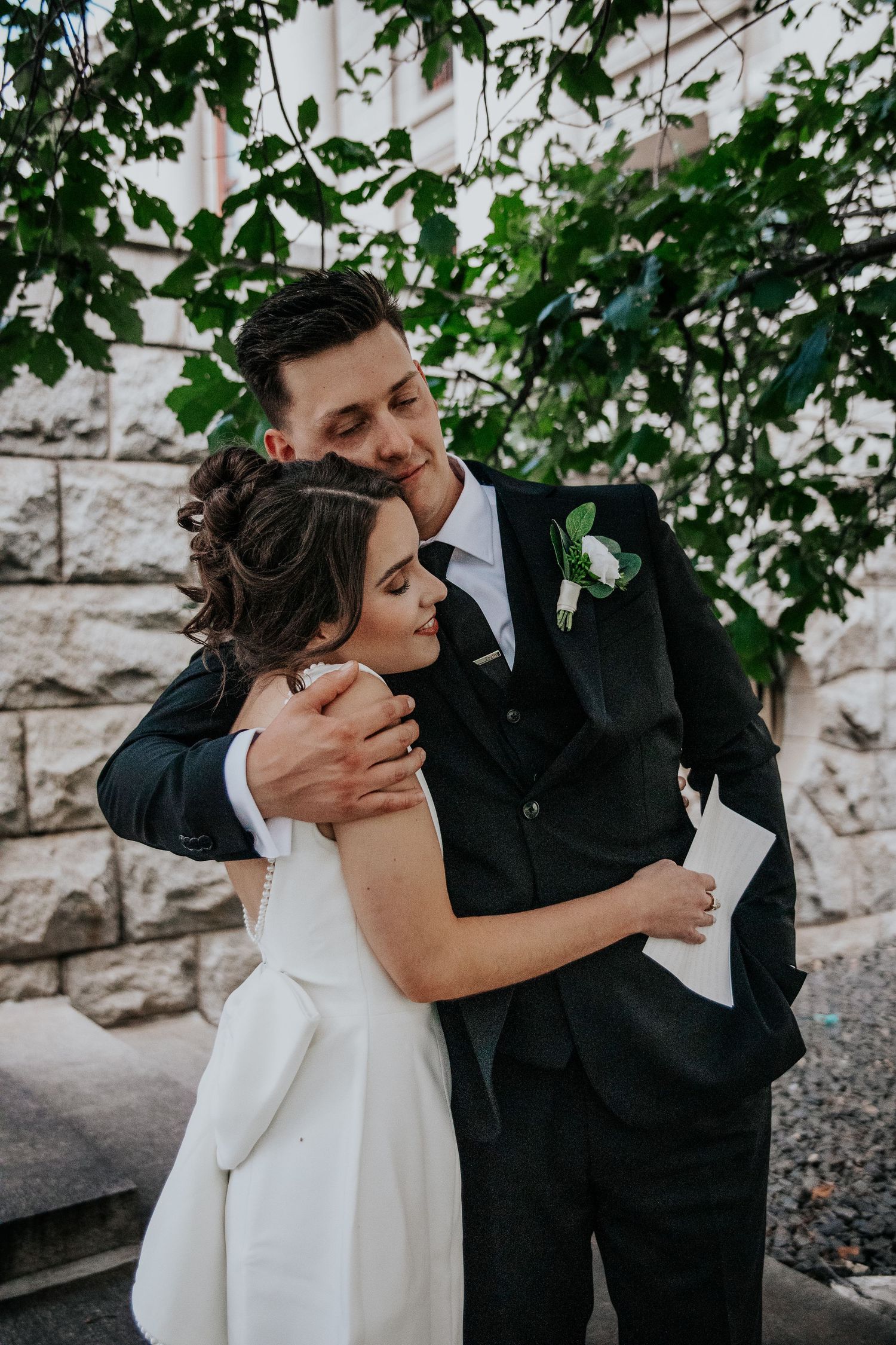 A romantic embrace between newlyweds under leafy green branches near a stone wall at Colorado Springs courthouse wedding