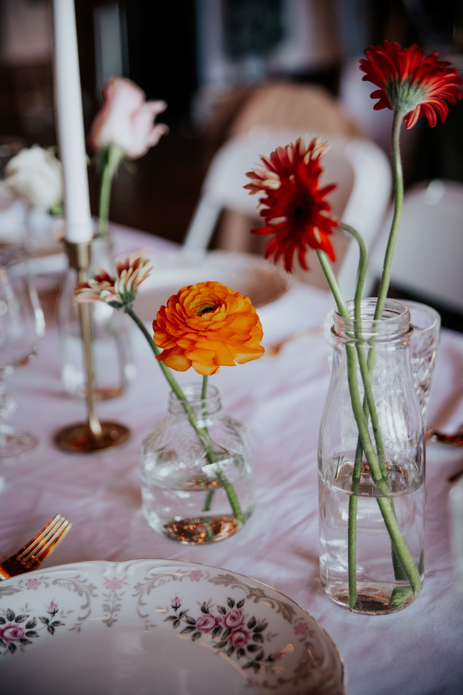Elegant table setting with red gerbera daisies and orange ranunculus in glass vases alongside floral patterned plates.
