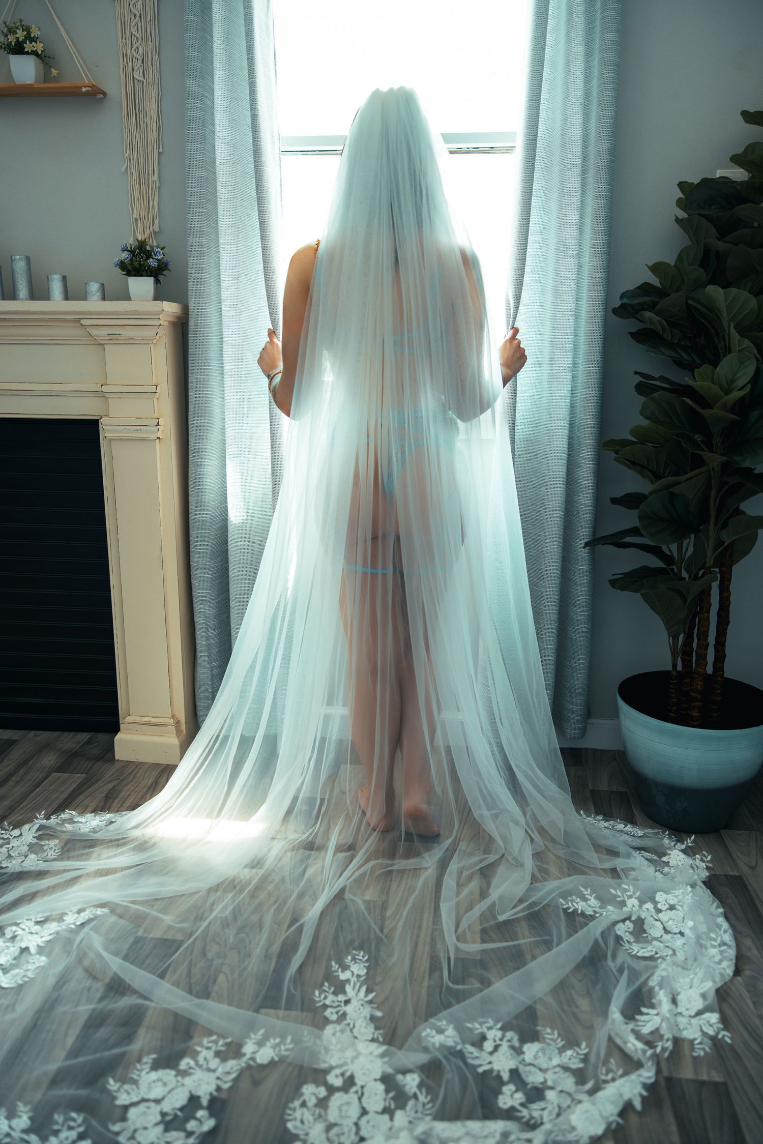 A bridal boudoir photo of a bride in teal lingerie with a  flowing cathedral-length wedding veil in standing by window with natural lighting