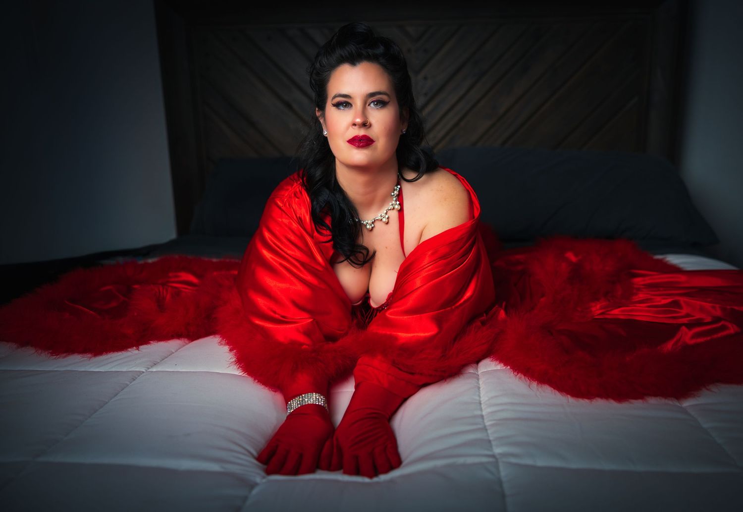 A dramatic boudoir photoshoot featuring a woman in red lingerie and red robe on a bed against dark and moody lighting.