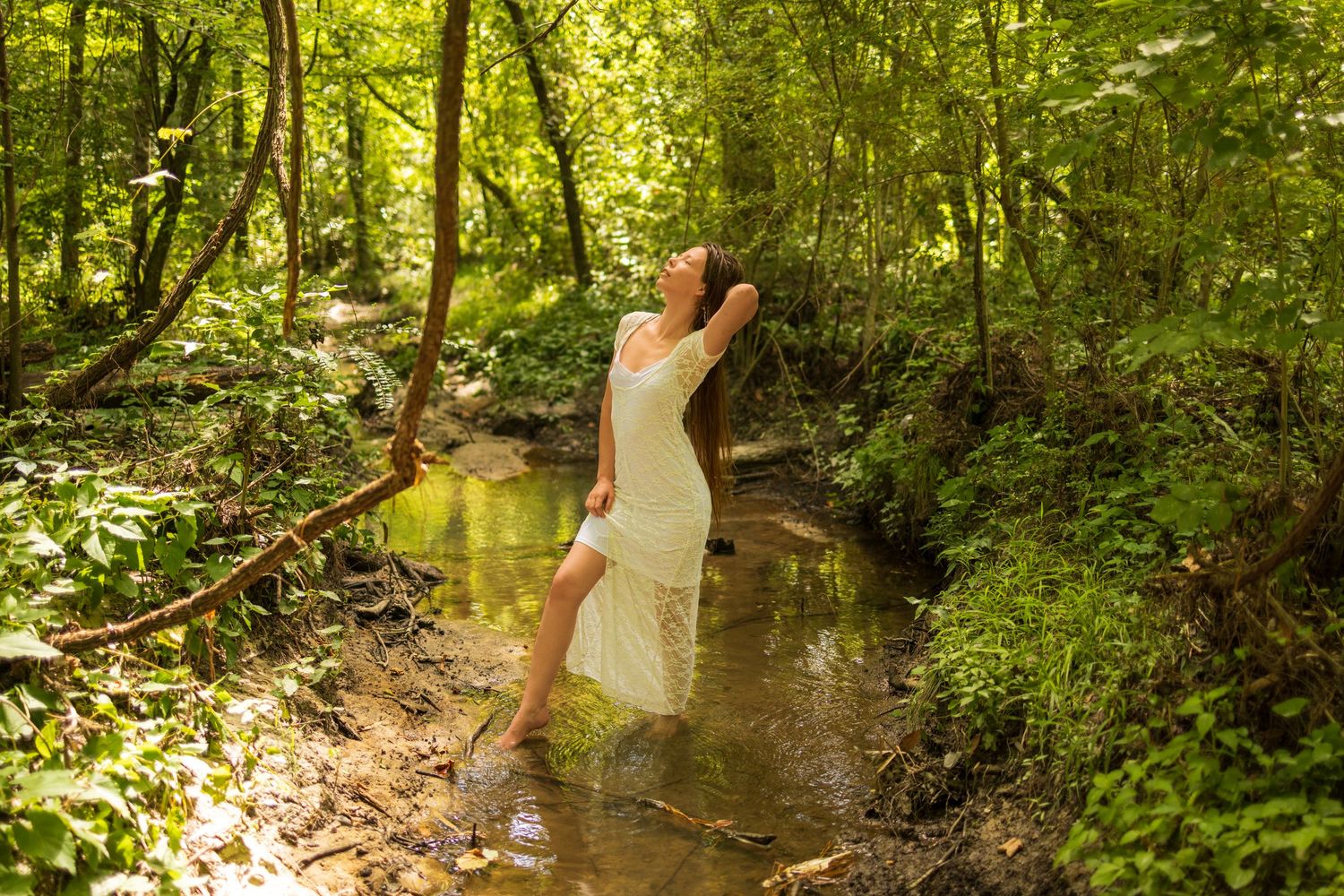 Artistic boudoir photo of a woman in white lace dress posed in a stream in a green lush forest