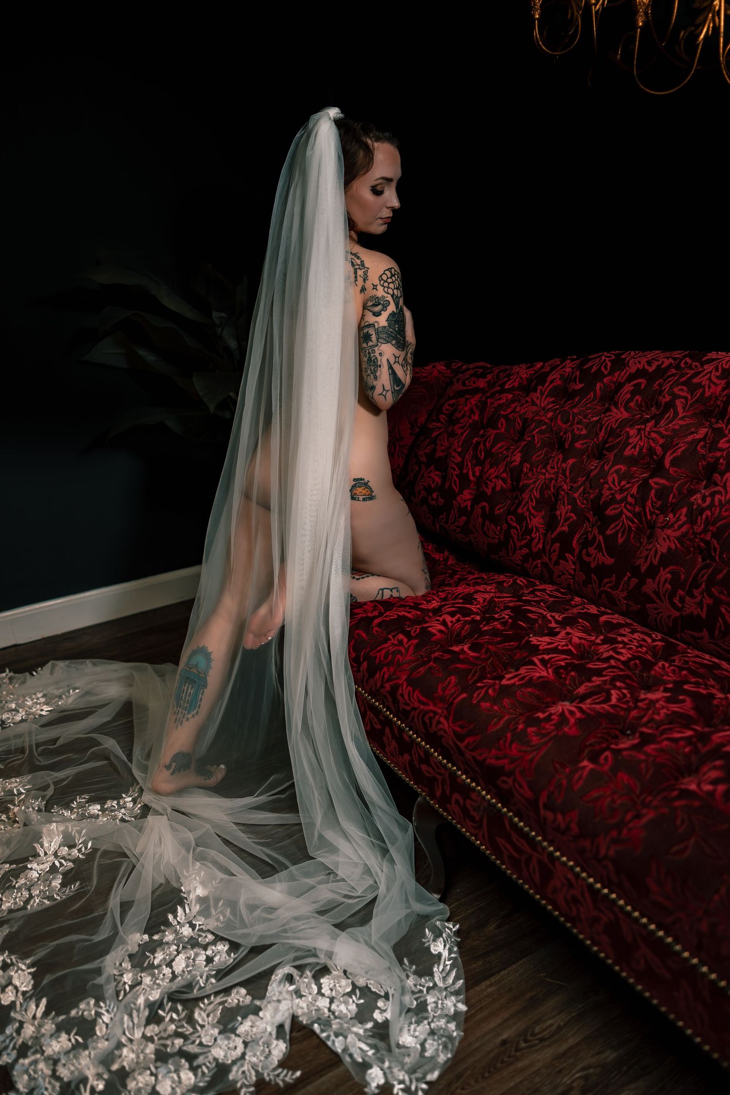 Artistic nude bridal boudoir portrait of a bride in a flowing white veil posed nude kneeling against a red velvet couch.