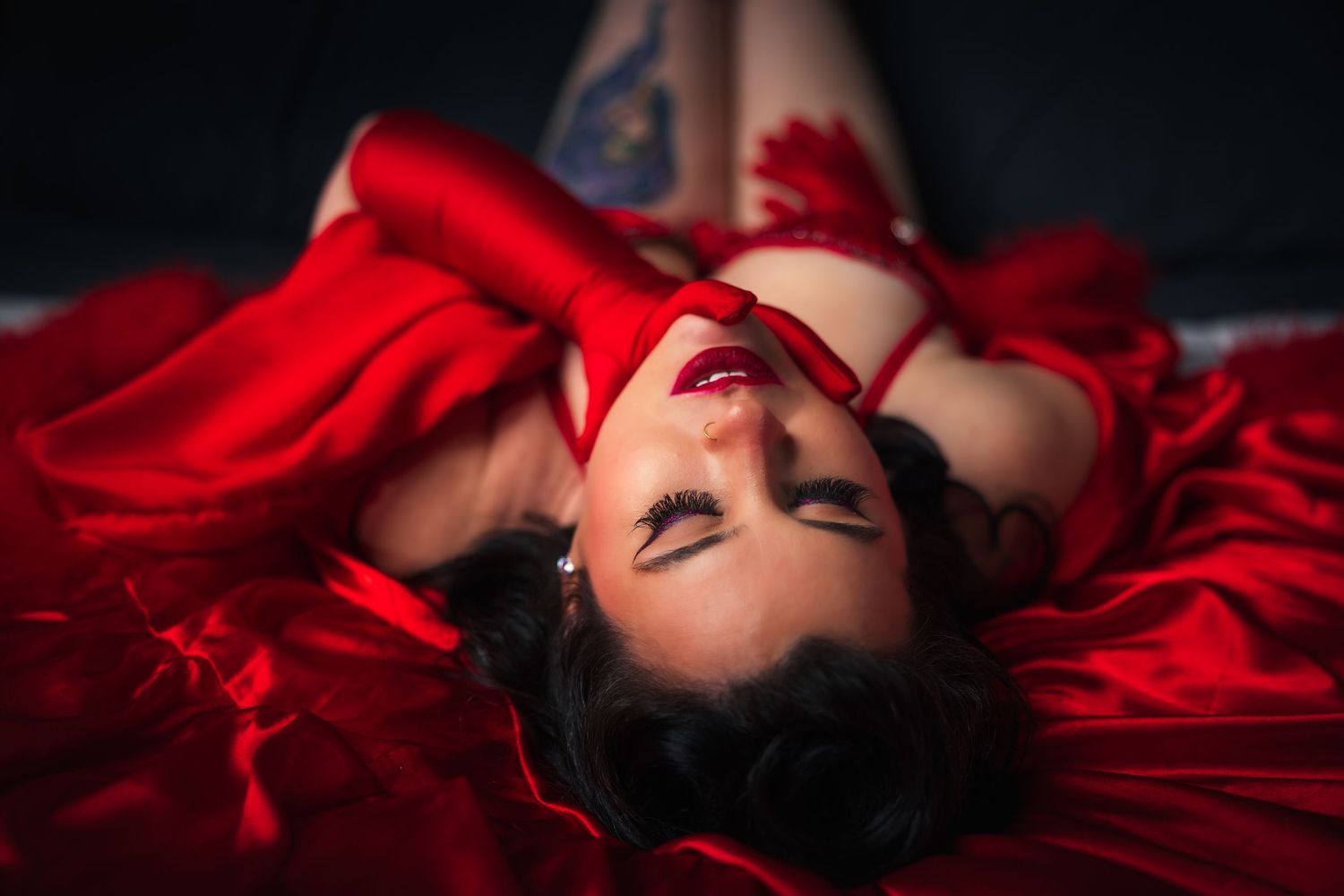 Person in dramatic lighting poses on red silk fabric creating artistic portrait series.