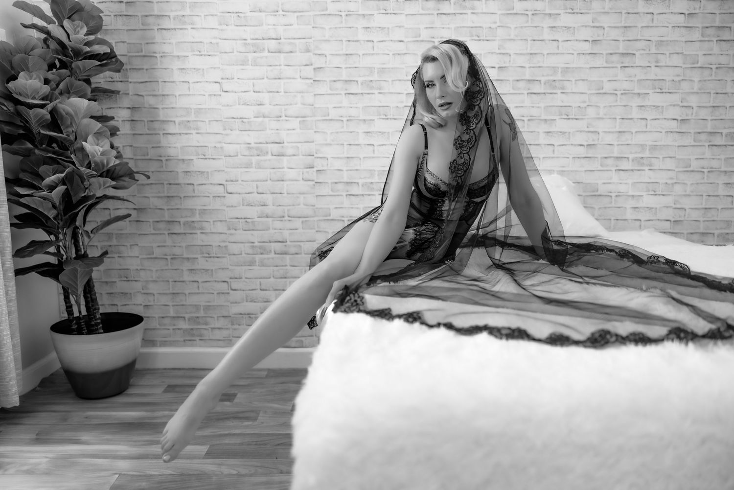 Black and white Bridal boudoir photo showing a woman in black bridal veil and black lingerie on a white bed with white brick wall.