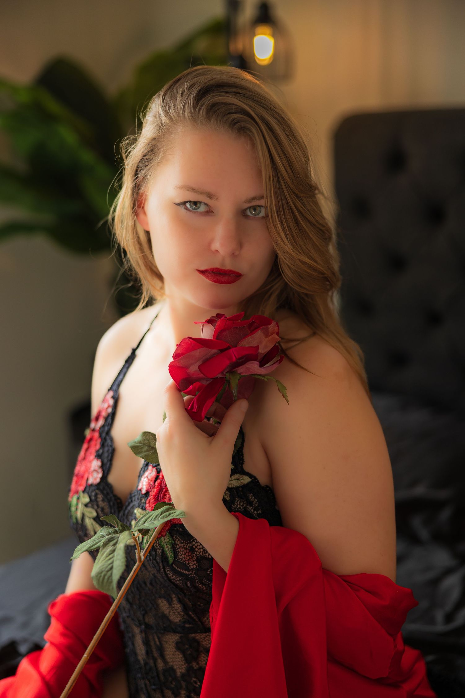 A romantic boudoir photo of a woman in black lingerie holding a red roses and red lace robe against soft lighting.