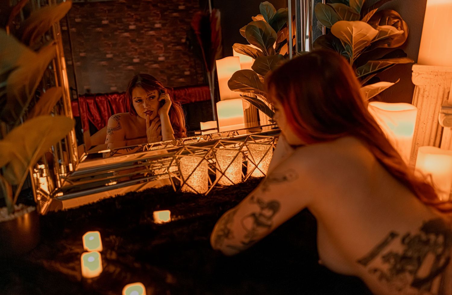 Artistic nude boudoir photo with woman in mirror, glowing candles creating moody atmosphere and warm lighting against dark background.