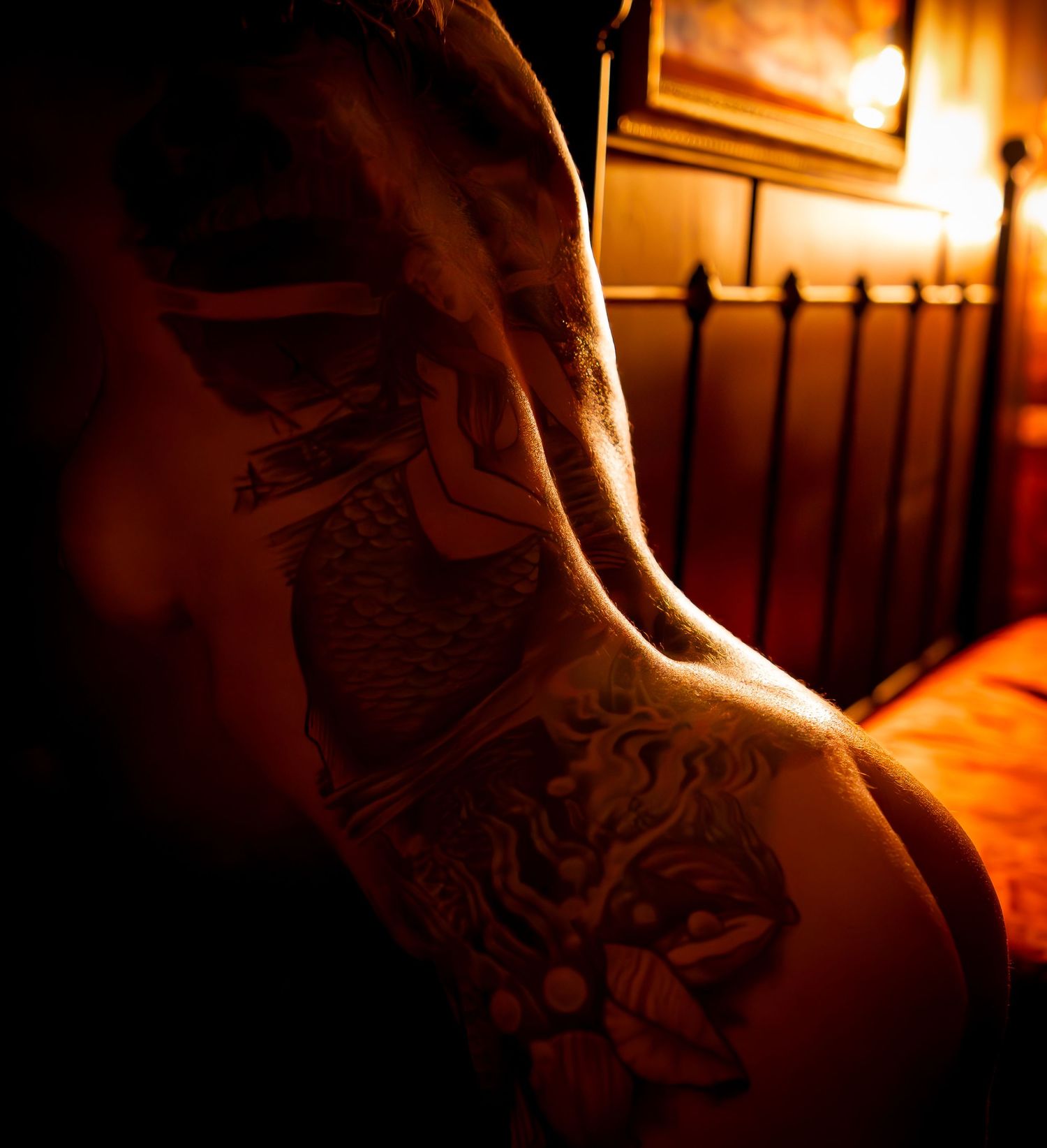 A artistic nude boudoir body scape detailed back side of woman illuminated by warm lighting in a dimly lit room with a bed frame visible.