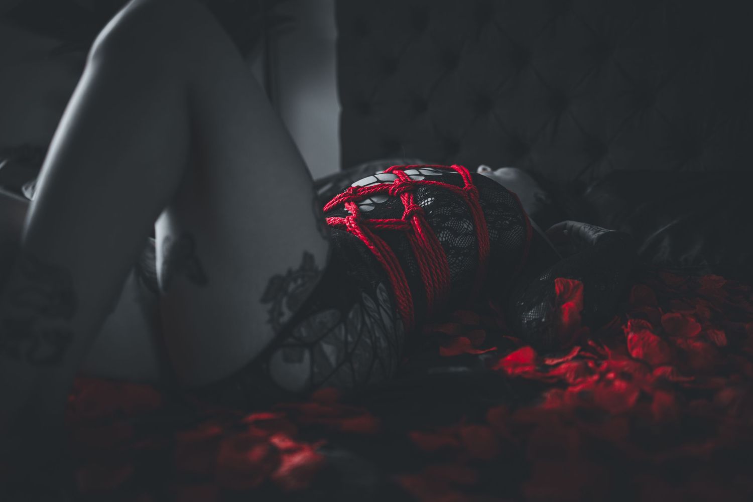 A black and white red colored photo of a woman tied in shibari rope bondage posed laying on a bed of red roses in black lingerie.