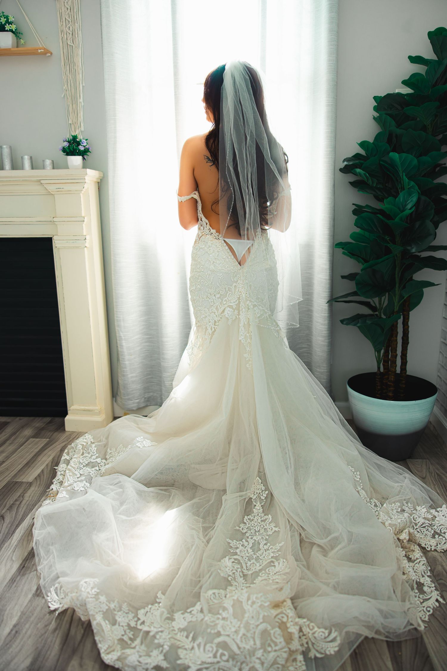 artistic bridal boudoir photo of woman in a white lace wedding gown with a dramatic train from behind in a sunlit room with a fireplace.