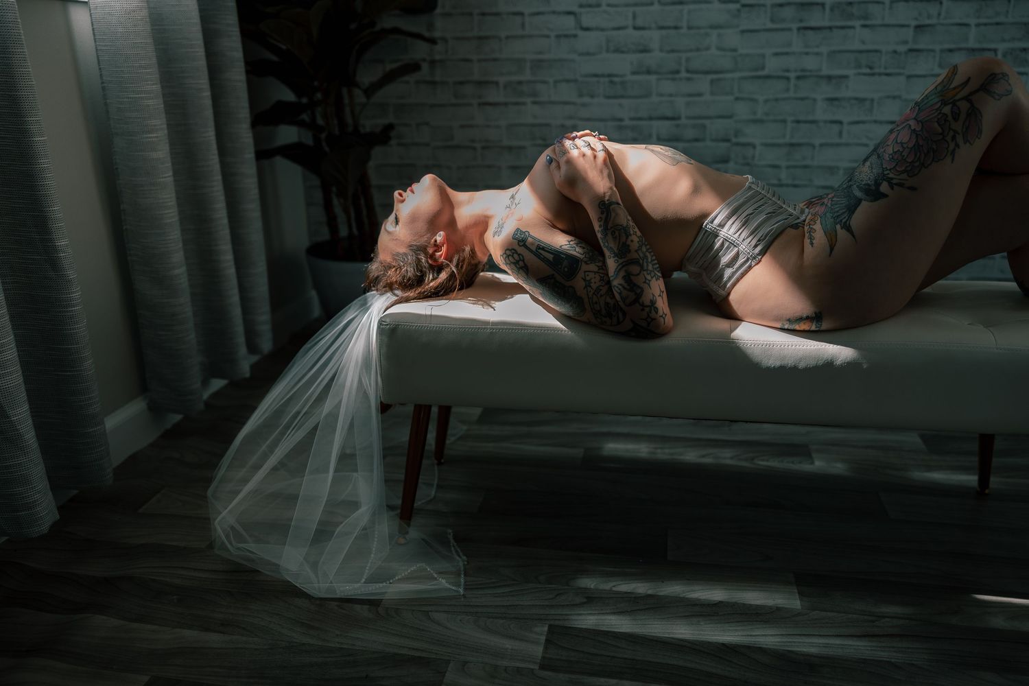 A dramatic bridal boudoir boudoir photo in moody lighting shows a figure reclining on a bench with flowing white veil in a dark room.