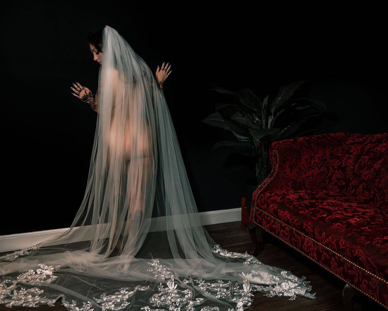 Artistic nude bridal boudoir portrait of a bride in a flowing white veil posed nude standing by a wall with a red velvet couch.