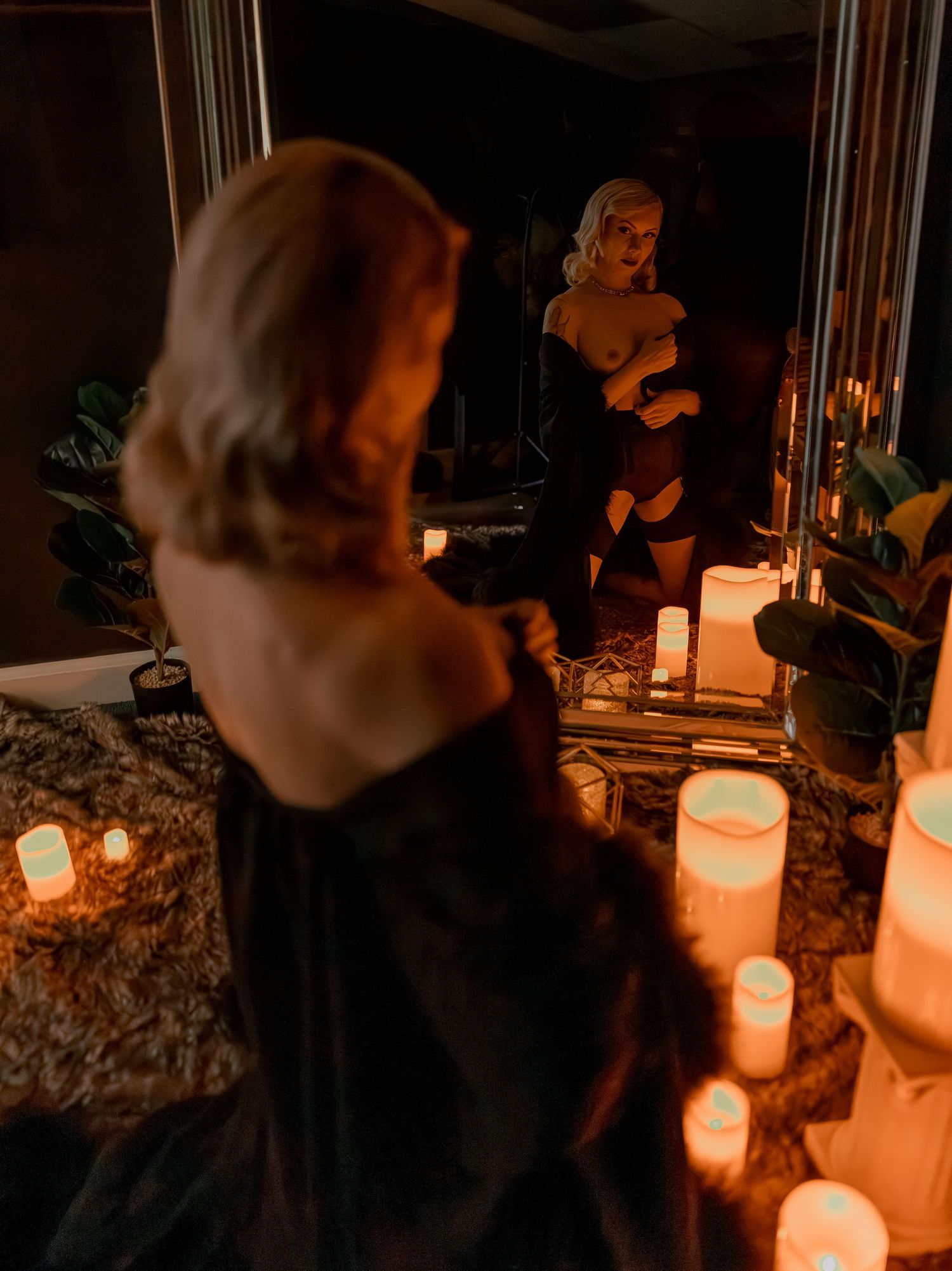 Artistic boudoir photo of a woman posed in black lingerie in front of mirror and glowing candles in a dimly lit room at night.
