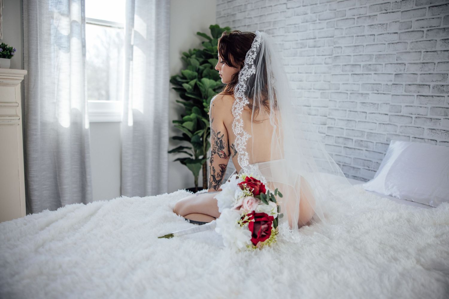 A artistic bridal boudoir photo of a woman in white lingerie and lace veil on white bedding with a red and white rose bouquet .