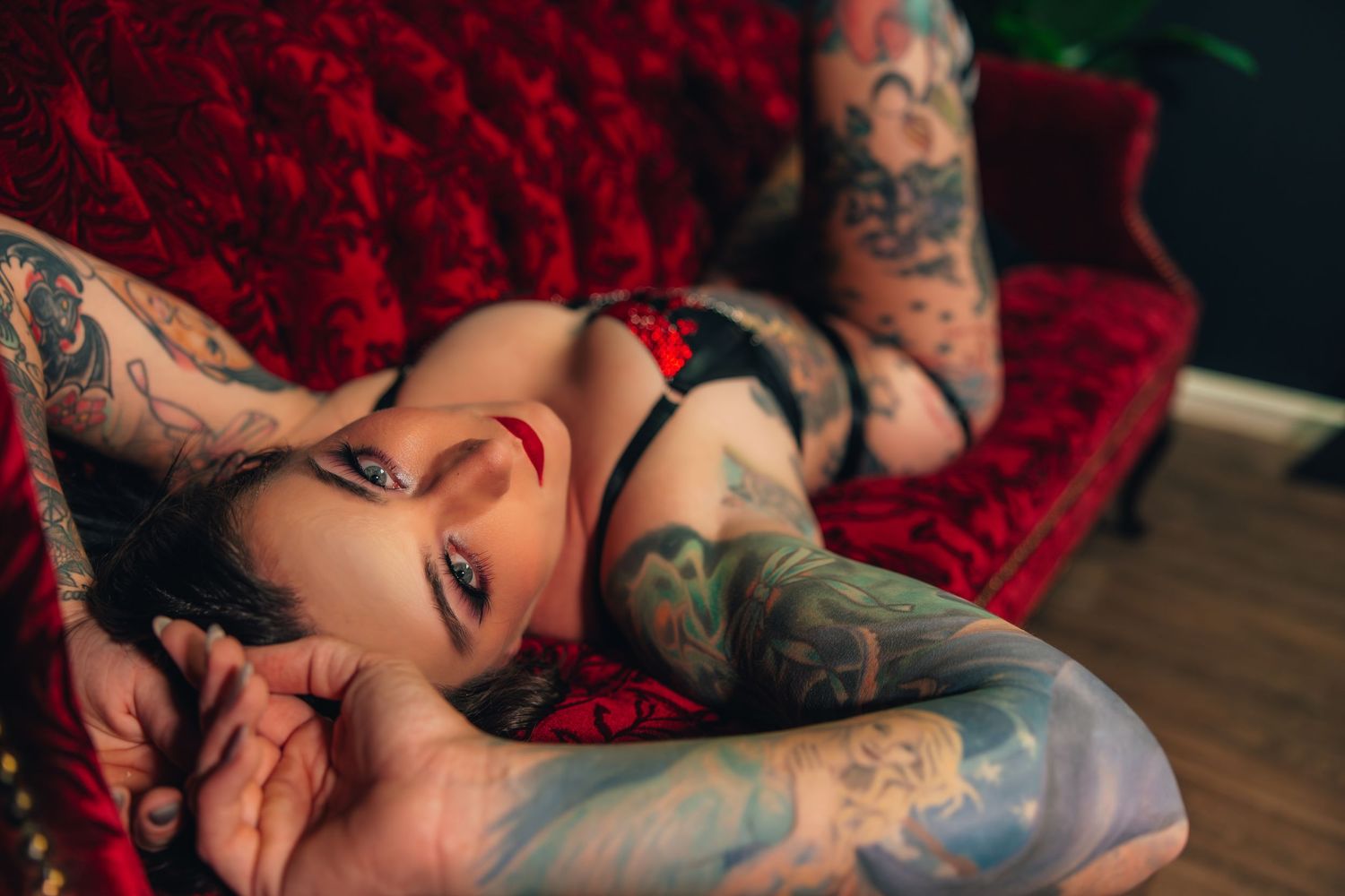 A boudoir photo of a woman with tattoos in black lingerie poses on a red velvet couch in an artistic boudoir photoshoot.