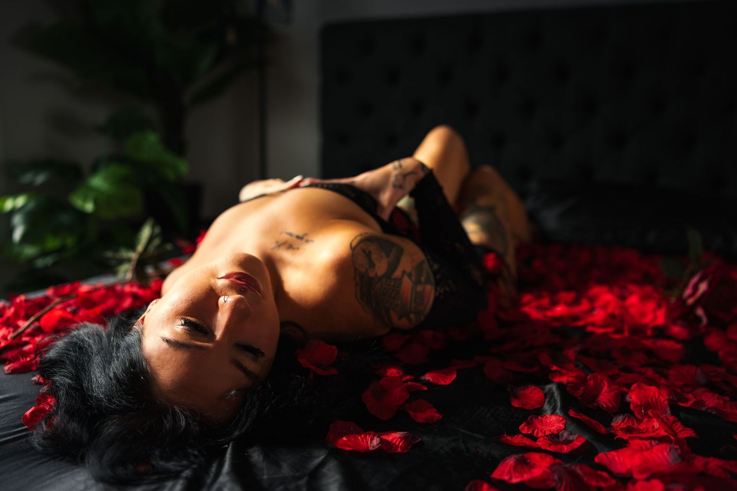 A boudoir photo of a woman in black lingerie lying on vibrant red rose petals in dramatic lighting creating a romantic image