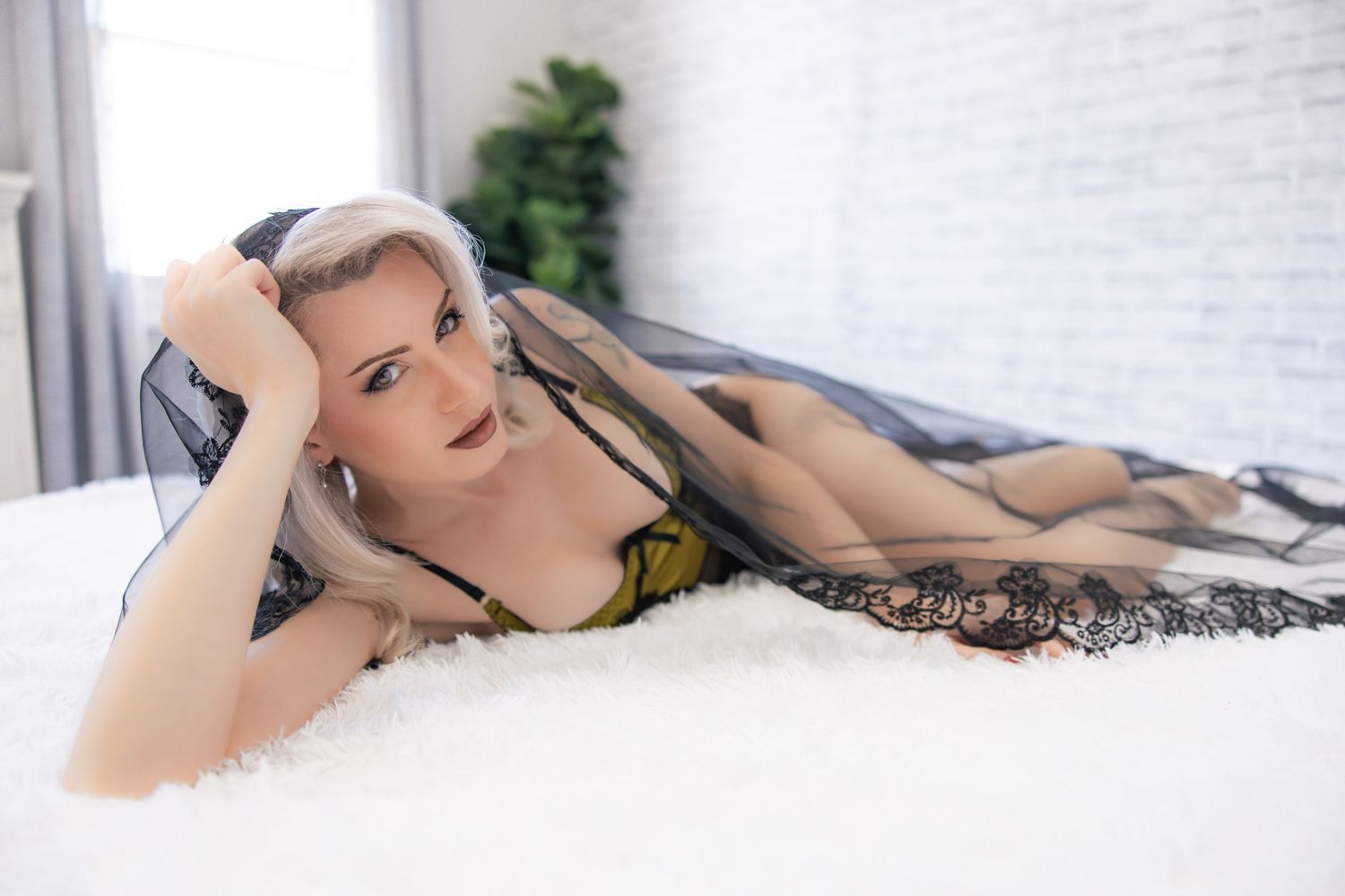 A Bridal boudoir photo showing a woman in black bridal veil and black and yellow lingerie on a white bed with white brick wall.