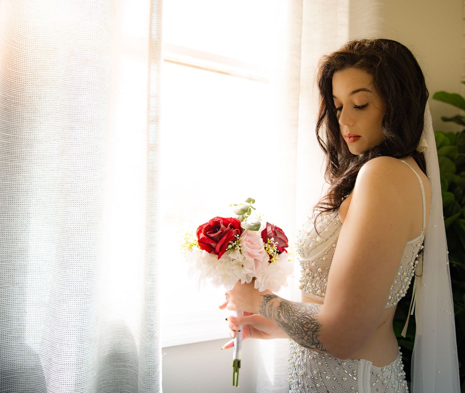 A bridal boudoir photo of bride in a white wedding dress holds a bouquet of red and pink roses near a window with soft natural light.