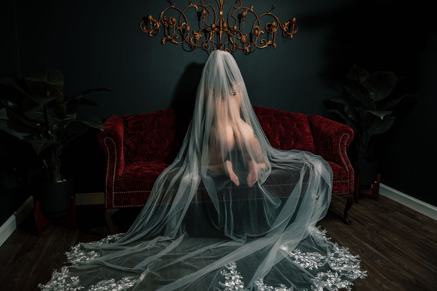 Artistic nude bridal boudoir portrait of a bride in a flowing white veil posed nude kneeling on a red velvet couch.