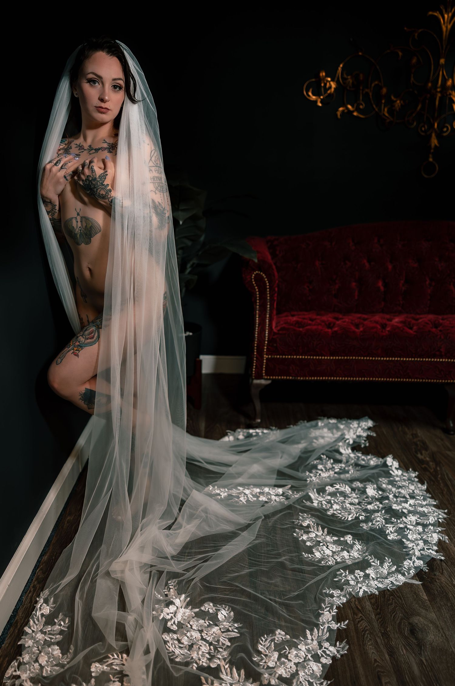 Artistic nude bridal boudoir portrait of a bride in a flowing white veil posed nude standing by a wall with a red velvet couch.