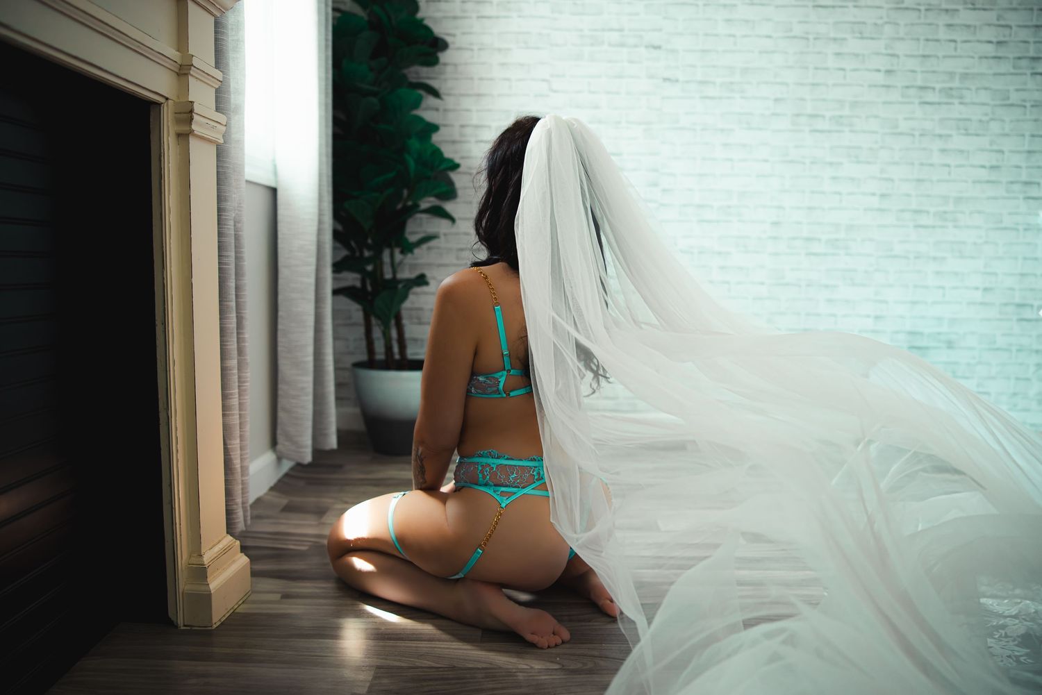 A bridal boudoir photo of a bride in teal lingerie and long flowing veil kneels on hardwood floor near white brick wall and fireplace.