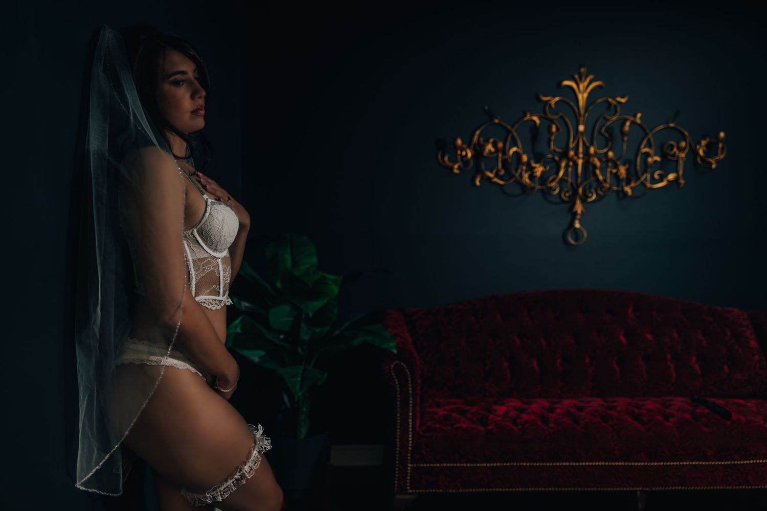 Artistic bridal boudoir photo of a woman in white lingerie with dark moody lighting and vintage red velvet furniture in elegant setting.