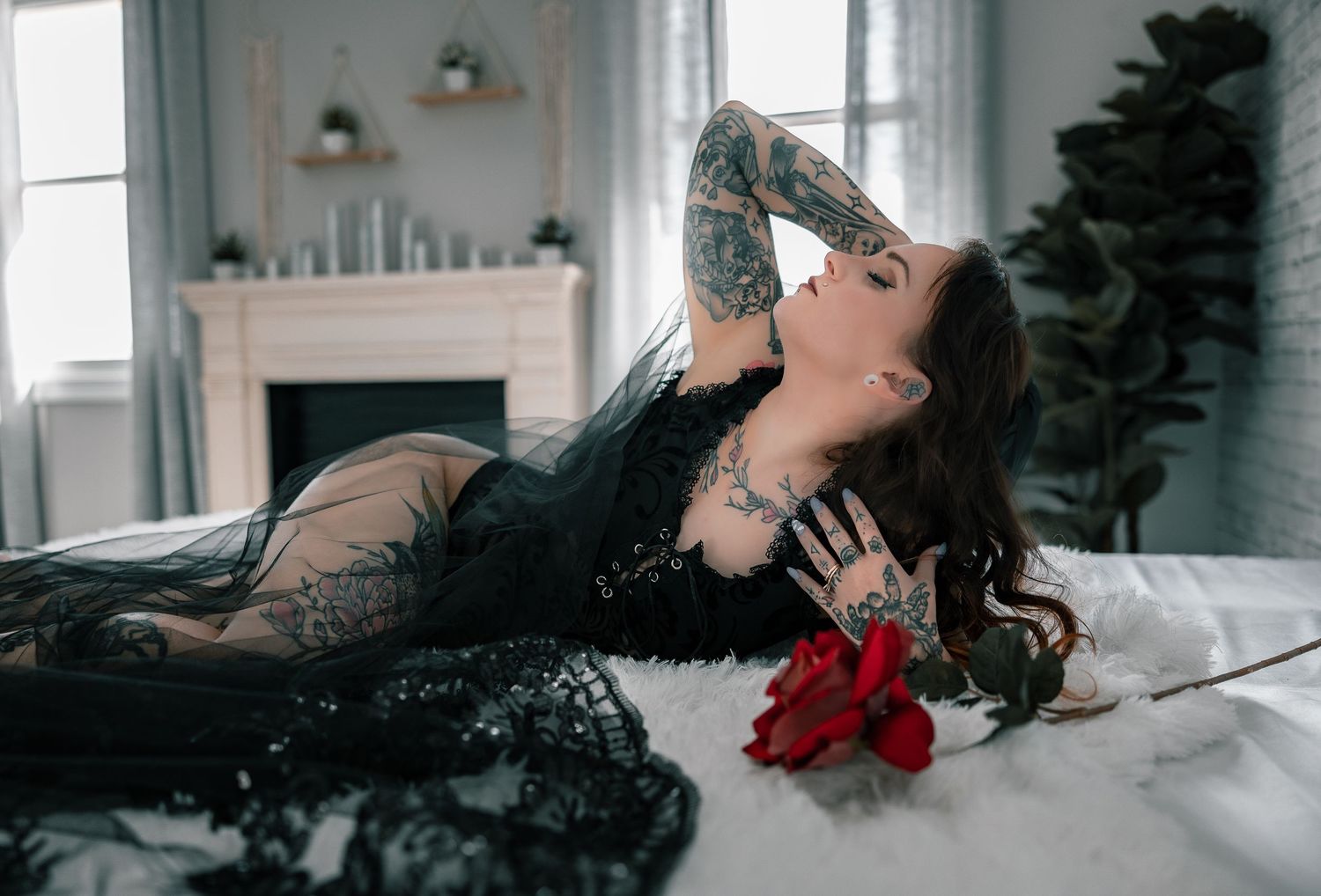 Artistic bridal boudoir portrait of woman in black lace dress and black veil lying on white bedding with red rose in moody indoor setting.