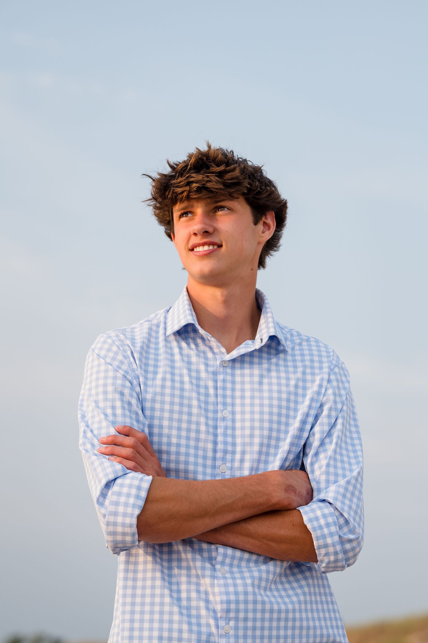 Senior guy in blue  shirt poses against light blue sky during beach photo session.