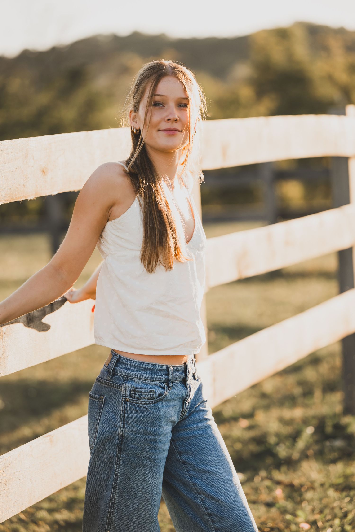 Senior girl  in white crop top and jeans poses casually against white fence in golden sunlight.