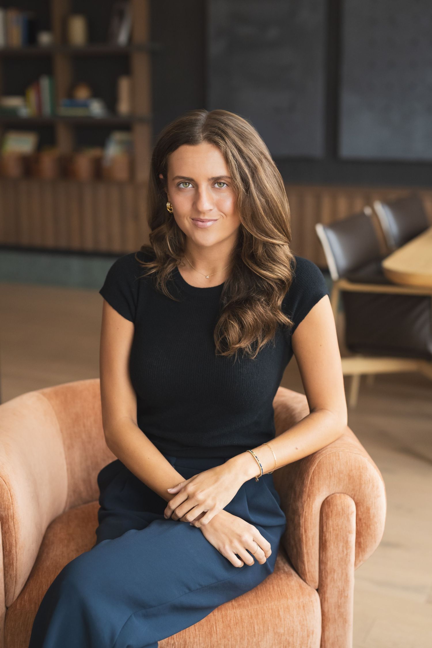 Woman in black top and navy skirt sits casually on a peach-colored armchair in a modern interior setting.