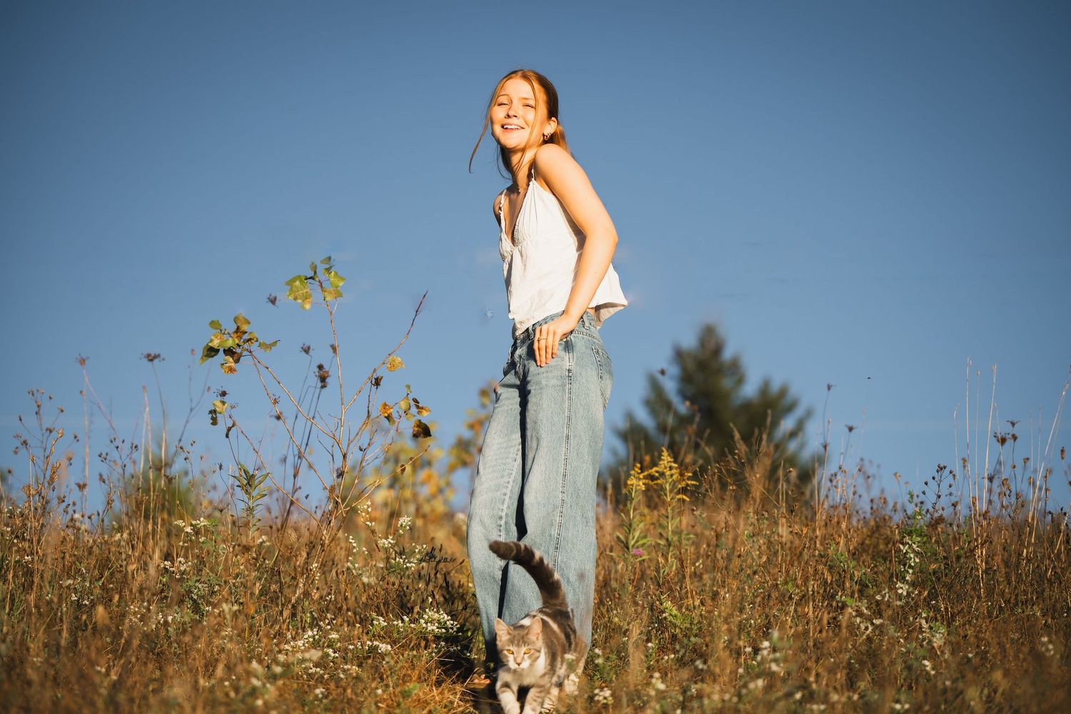 A person wearing a white tank top and jeans stands in a sunny meadow with a dog on a leash.