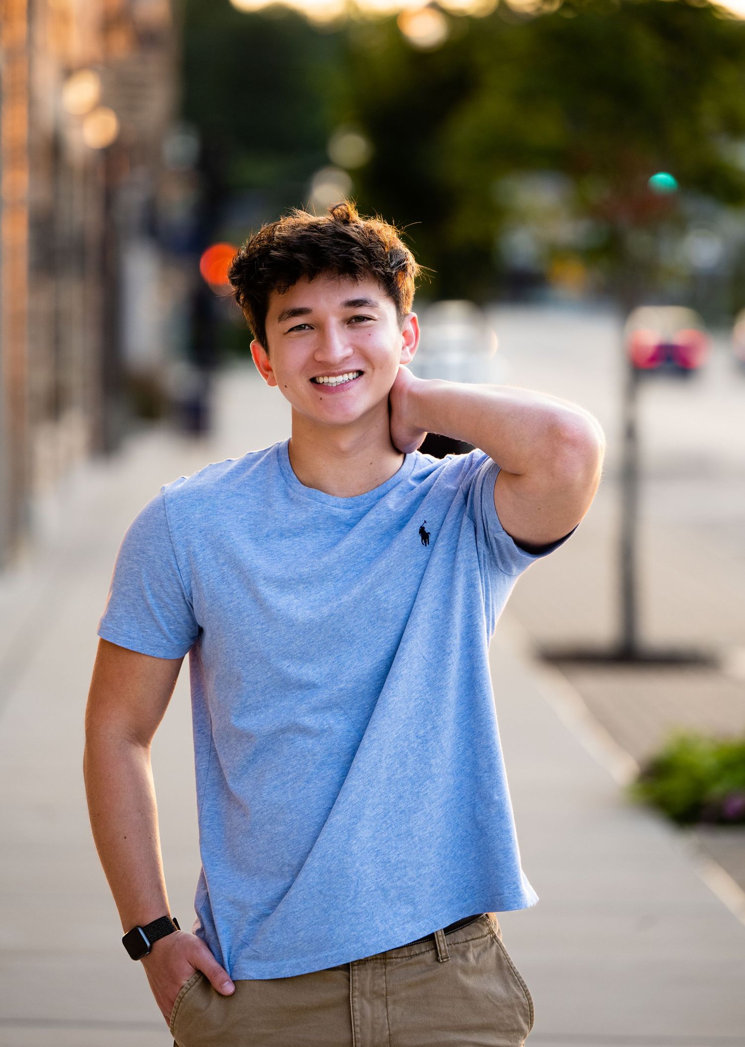 Person in light blue t-shirt and khaki shorts poses outdoors on city street during golden hour lighting.
