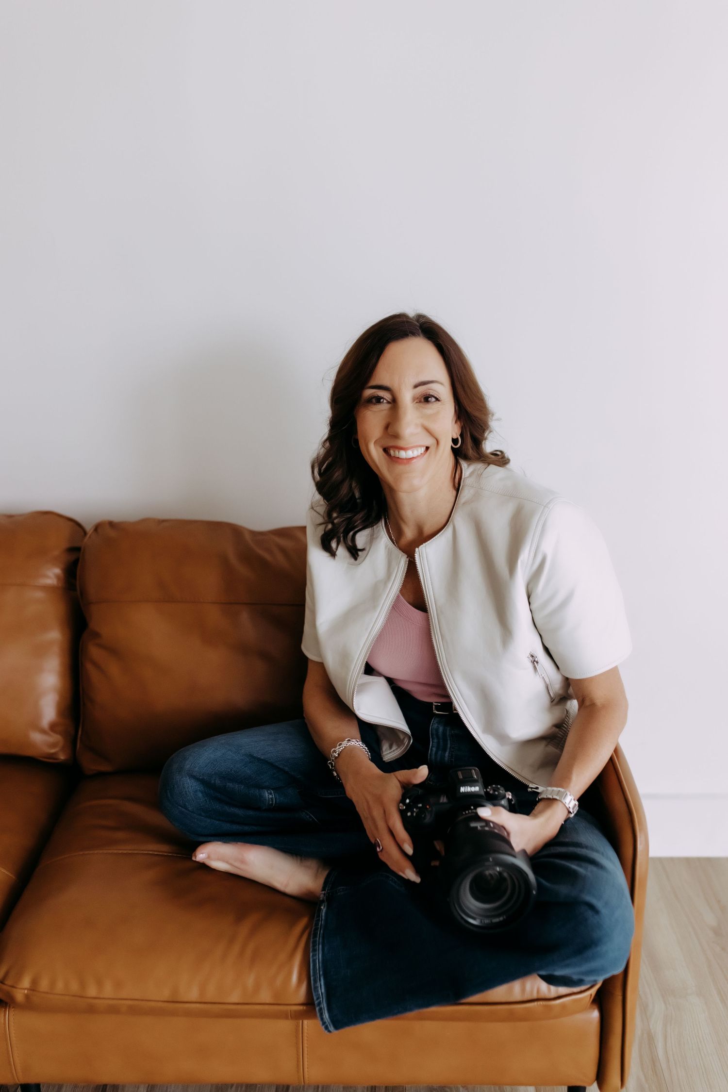 Woman photographer smiling on brown leather sofa holding DSLR camera, wearing white vest and jeans.