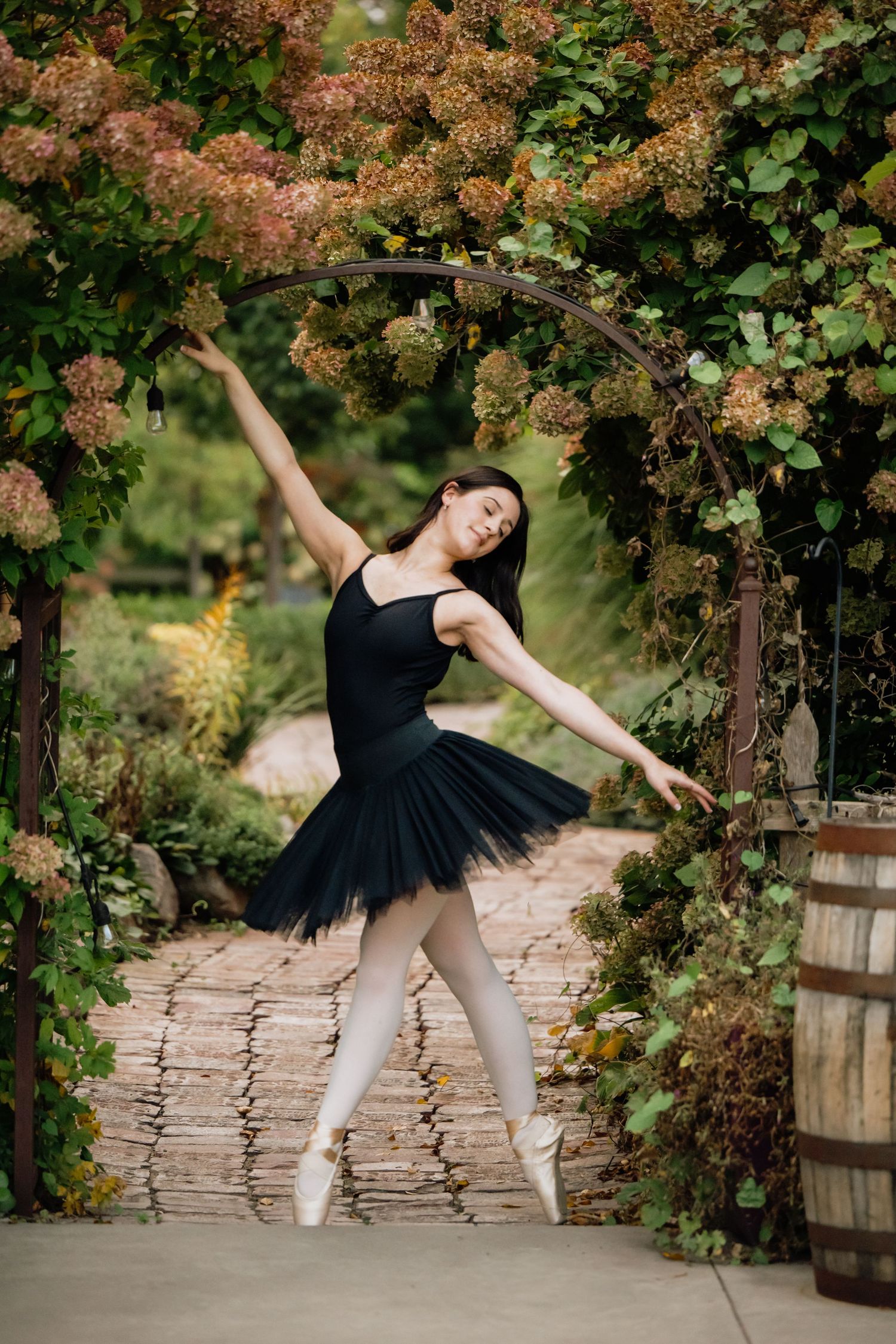 Graceful dancer in black tutu and pointe shoes performs elegant arabesque pose under flowering garden archway.