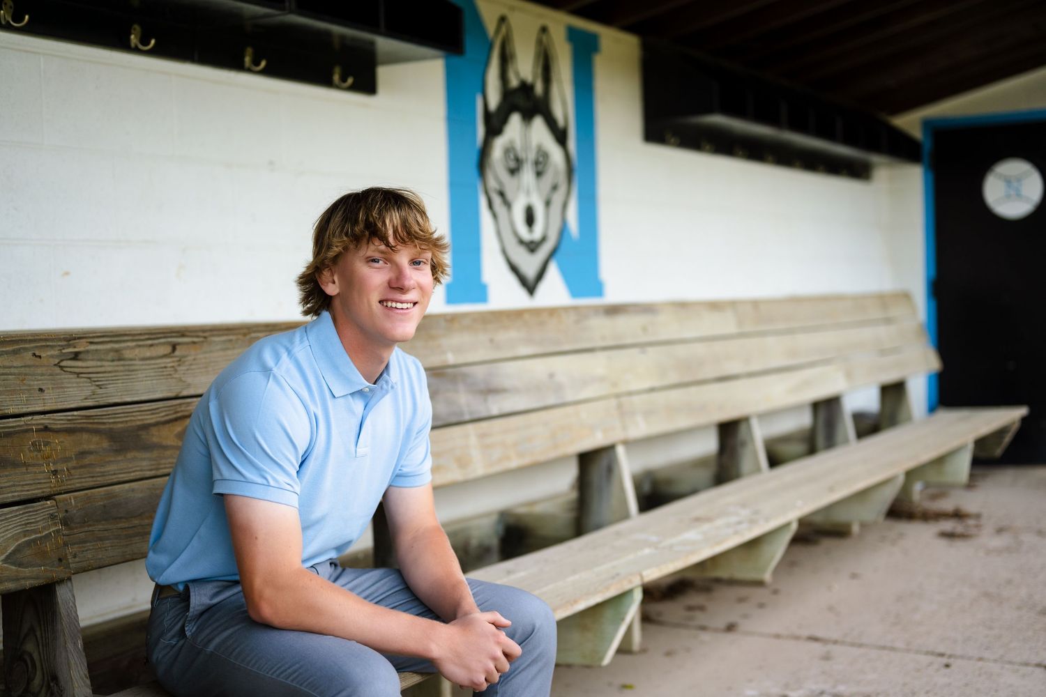 Senior boy on wooden bleachers in front of a wall decorated with a husky mascot logo.