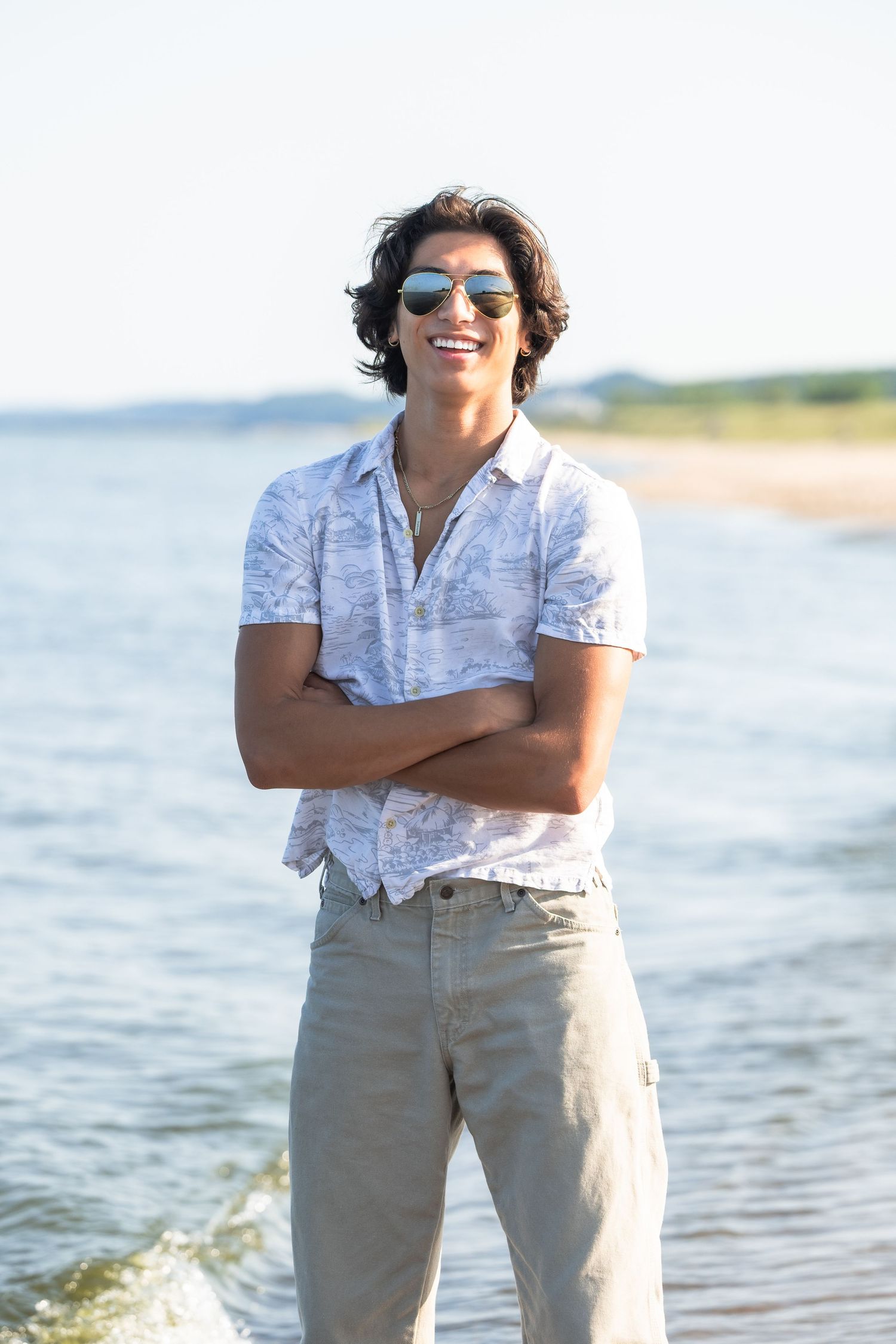 Smiling young man with sunglasses and crossed arms standing at a sunny beach wearing a patterned shirt and khaki pants.