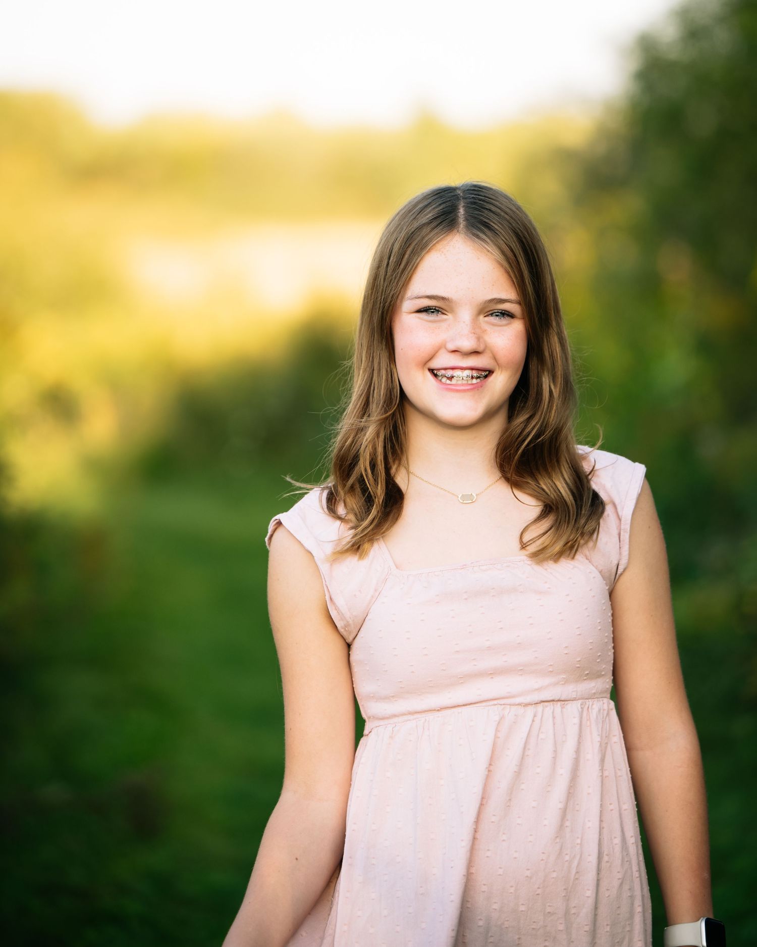 A girl in a light pink dress smiles warmly while standing outdoors against a soft blurred natural background.